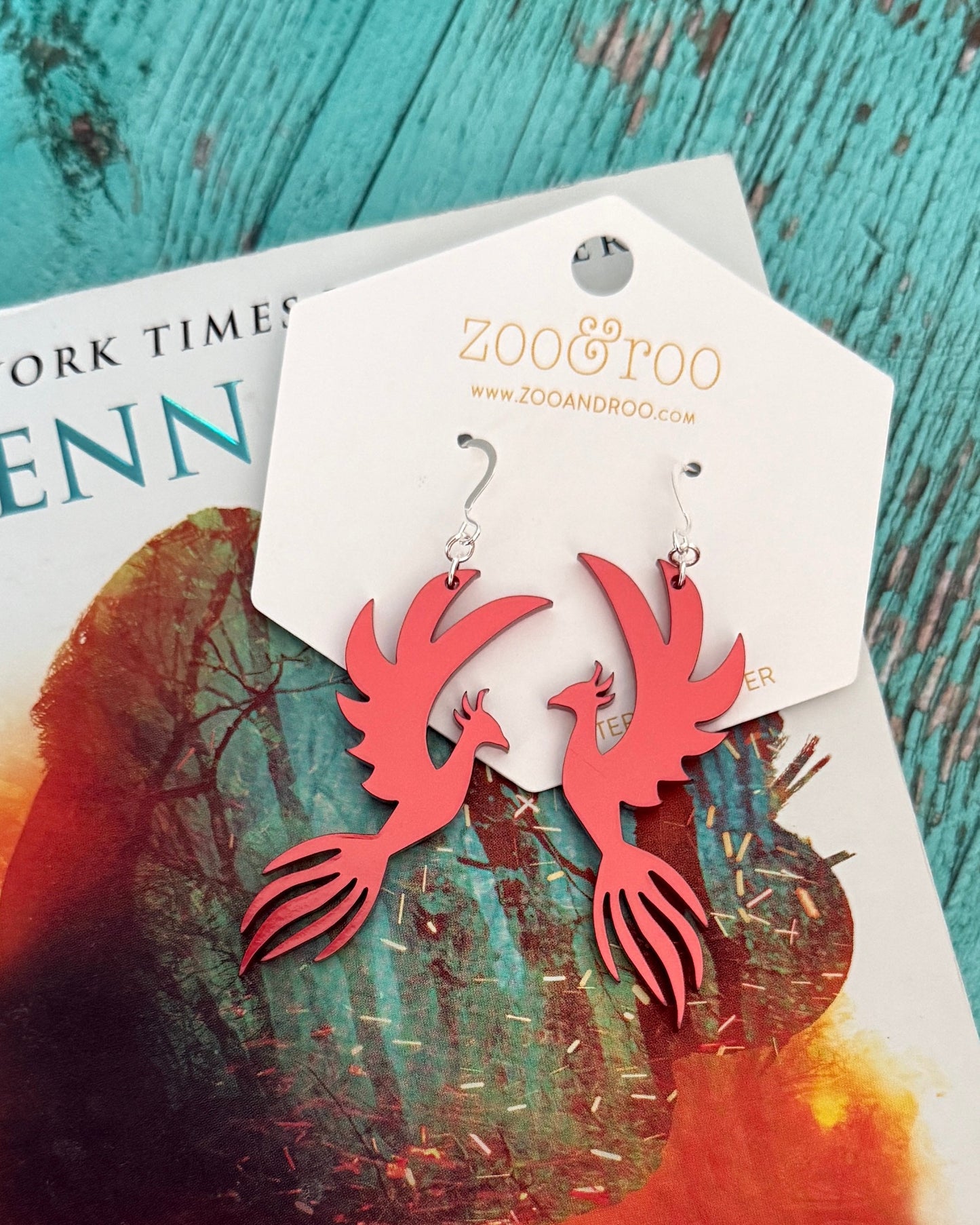 Red metallic phoenix earrings on a zoo&roo branded card laying on the Spark of the Everflame book by Penn Cole and a teal wood background