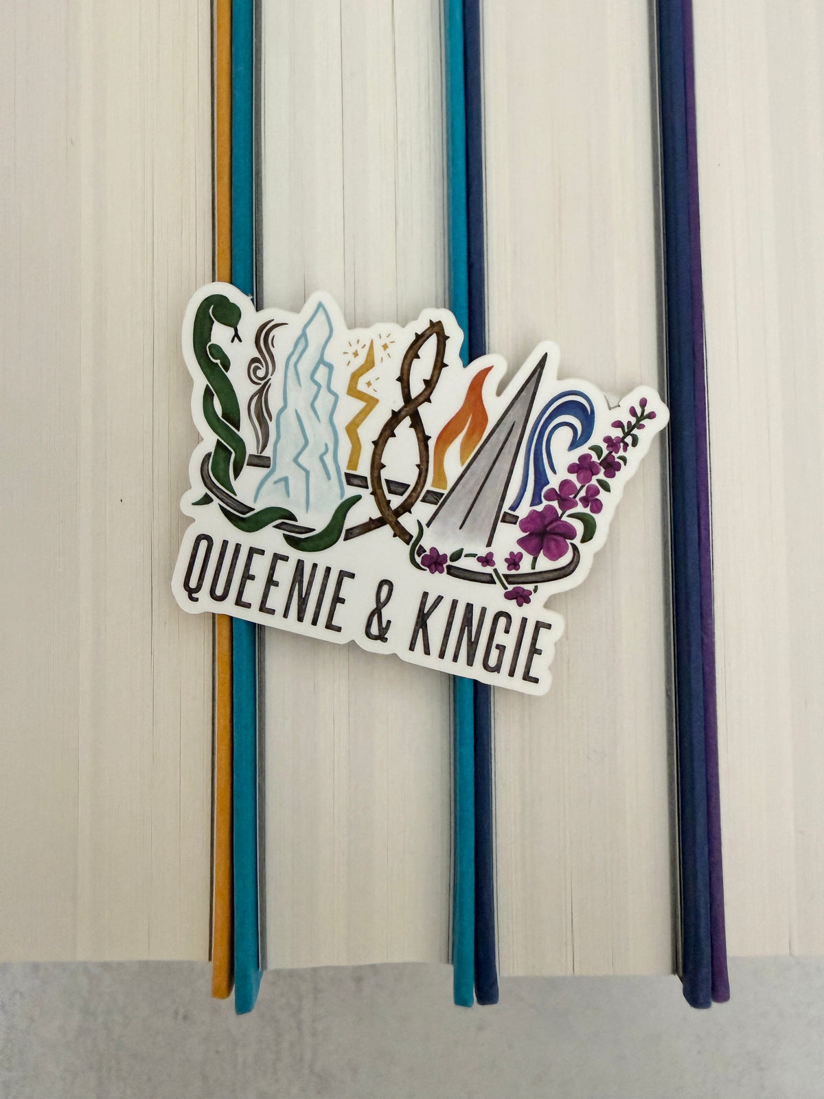 A sticker of a nine pointed crown featuring all the realms of Emarion from the Kindred's Curse Saga books by Penn Cole, with the text 'Queenie & Kingie' laying atop colorful books.