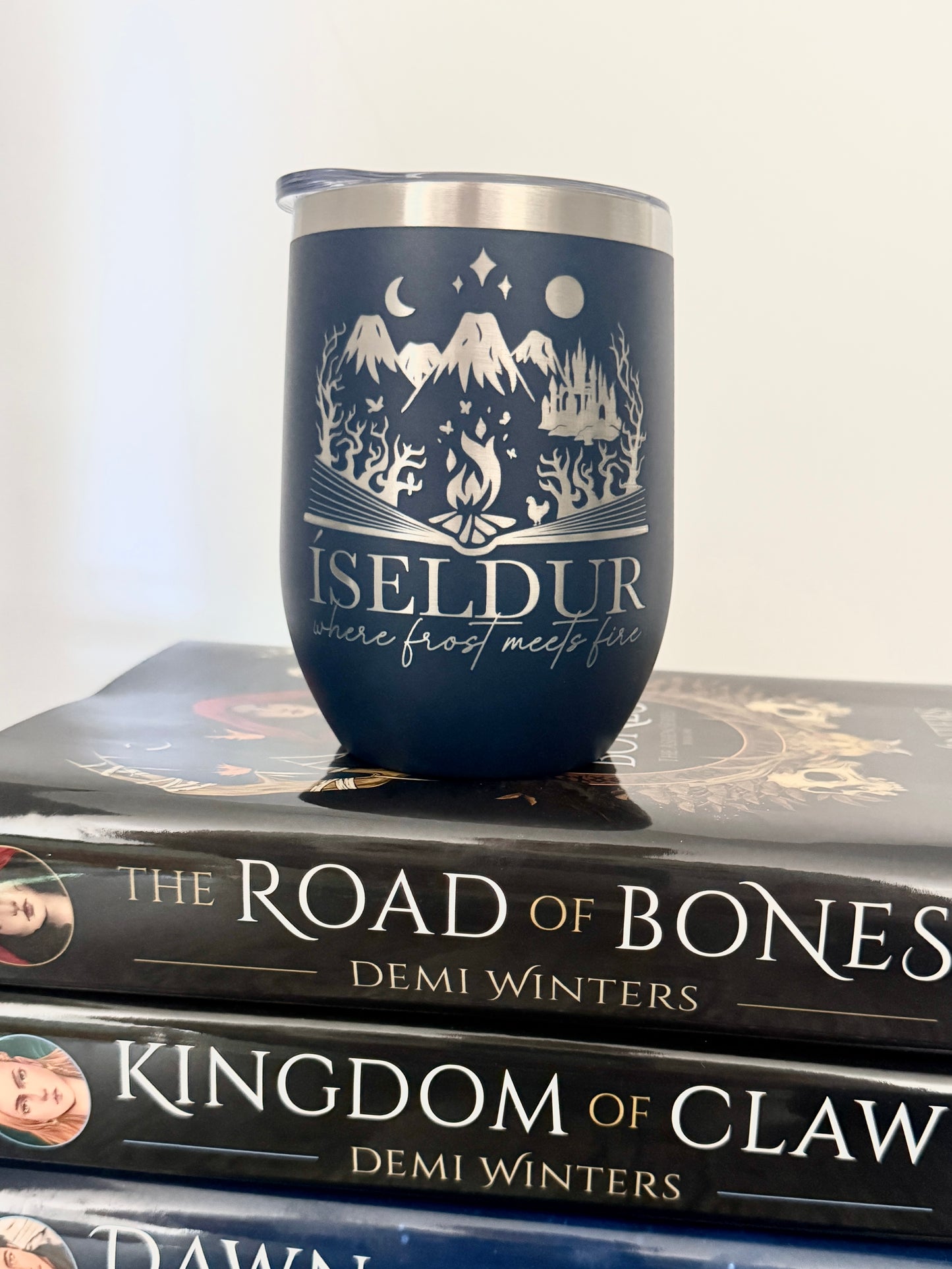 Navy blue wine tumbler with fantasy mountain design and 'Iseldur: where frost meets fire' text on a stack of Demi Winters' Ashen series books