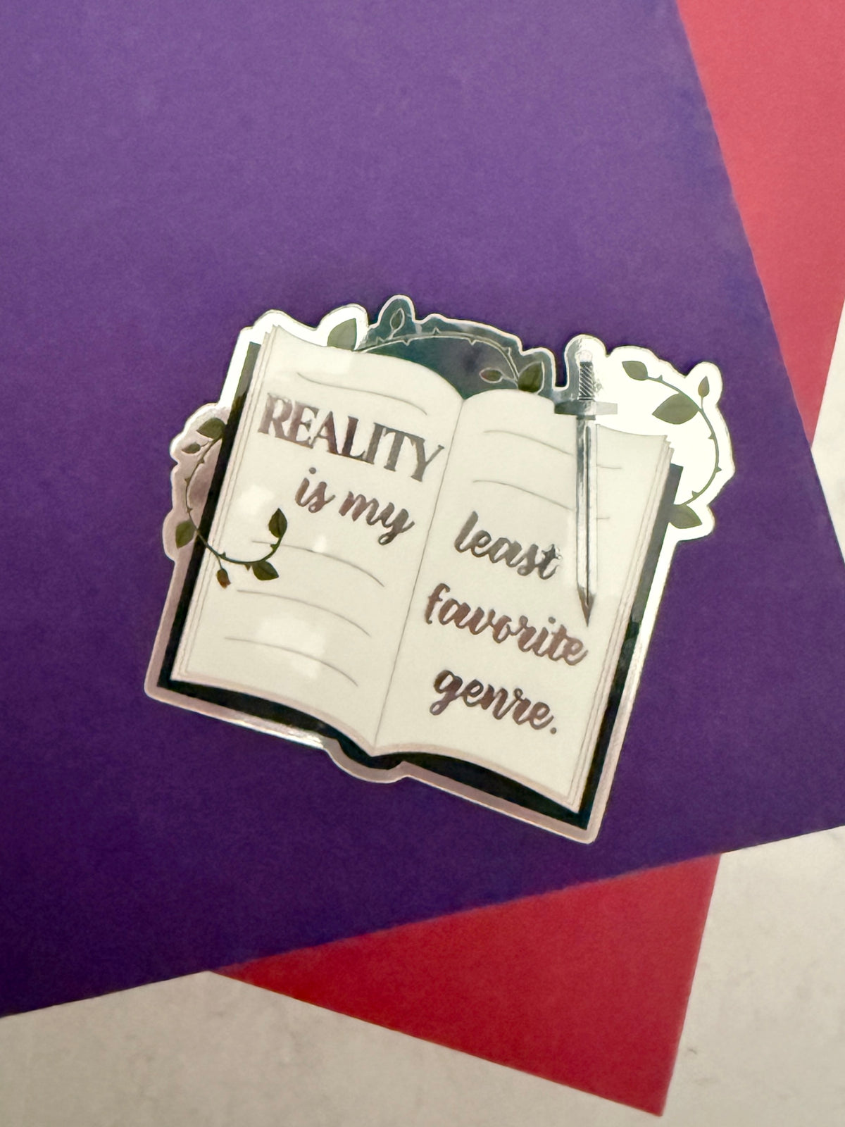 Open book sticker with metallic lettering 'reality is my least favorite genre,' a sword, and thorned vines, sitting on top of purple and red books