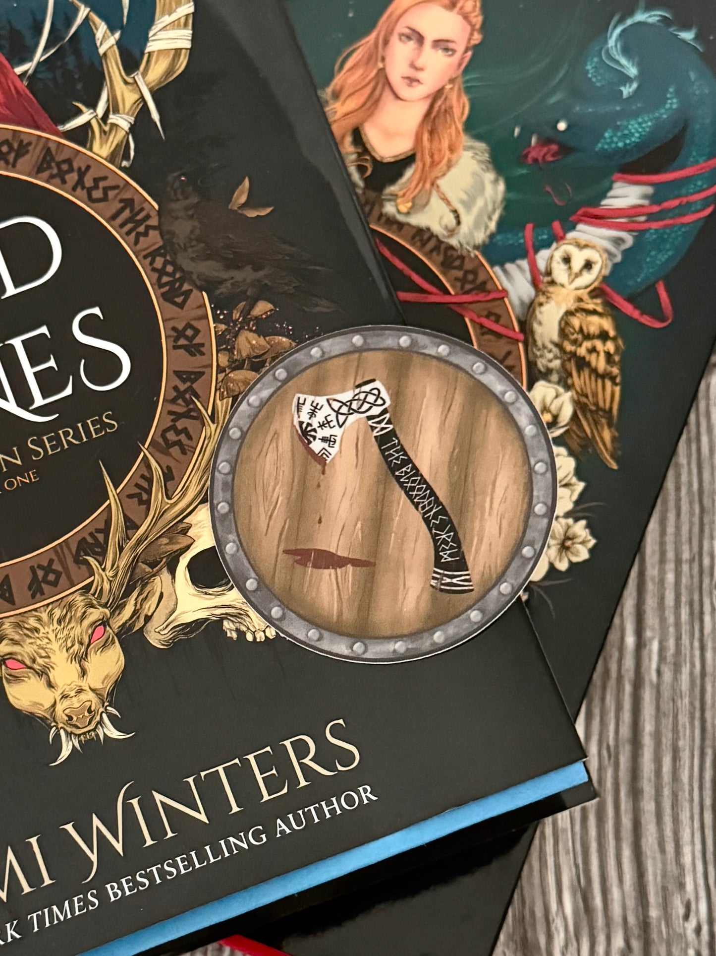 Round viking shield sticker with an axe drawn on it that reads The Bloodaxe Crew in runes, laying on The Road of Bones and Kingdom of Claw books by Demi Winters.