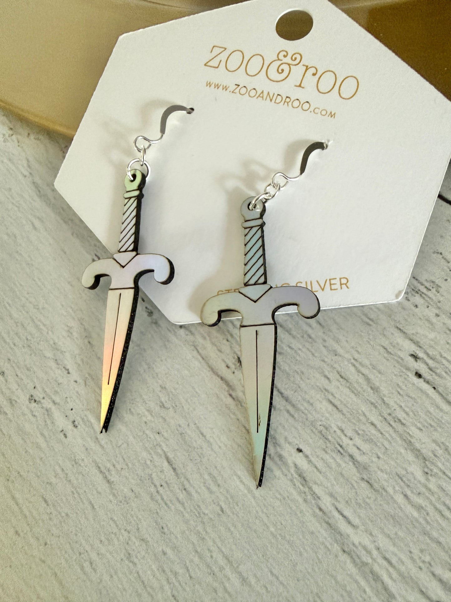 A pair of iridescent vegan leather dagger earrings, displayed on a white zoo&roo brand card against a light gray wood background.