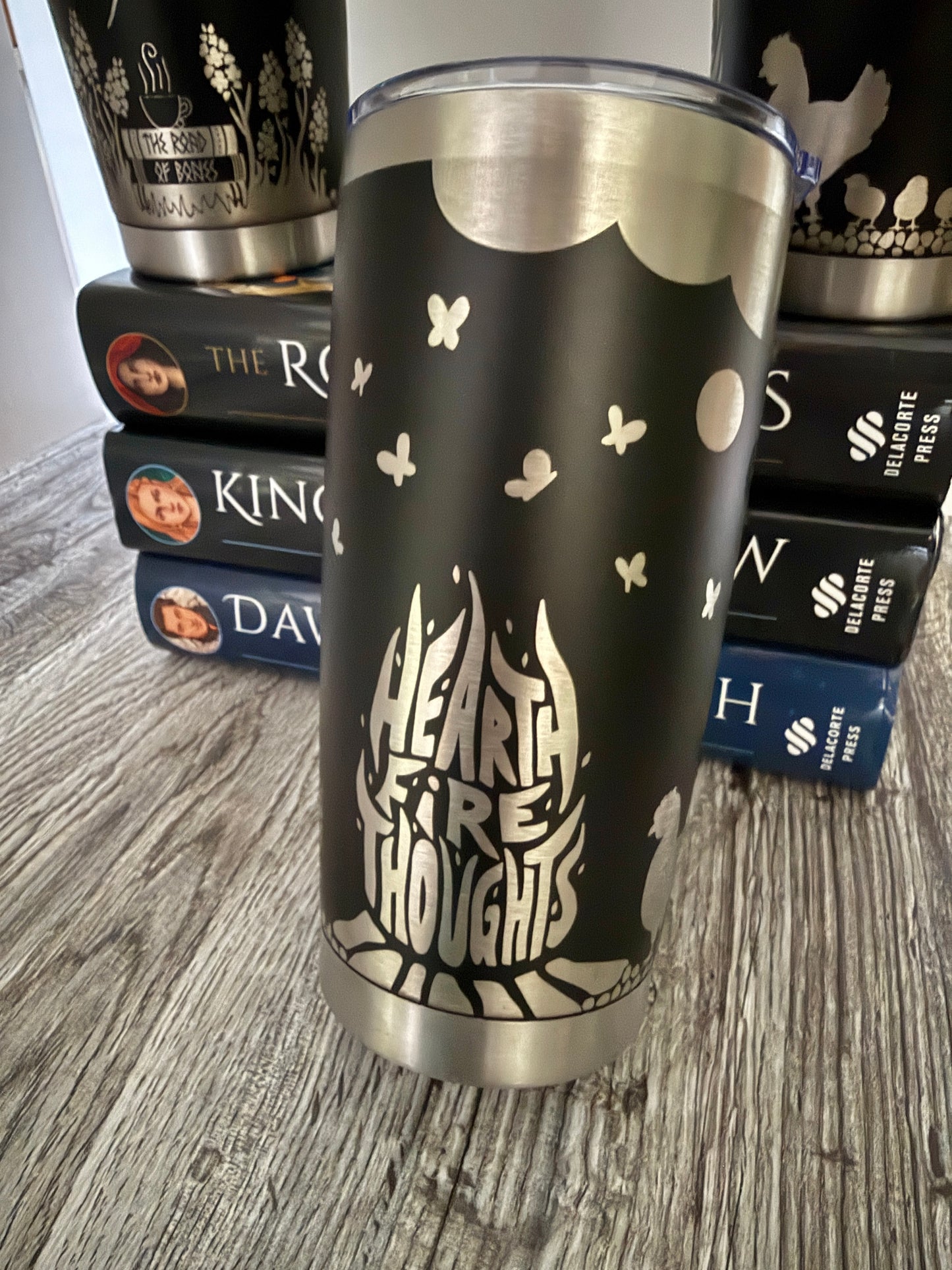 Black tumbler with 'Hearthfire Thoughts' design on a wooden surface with Demi Winters' Ashen series books in the background