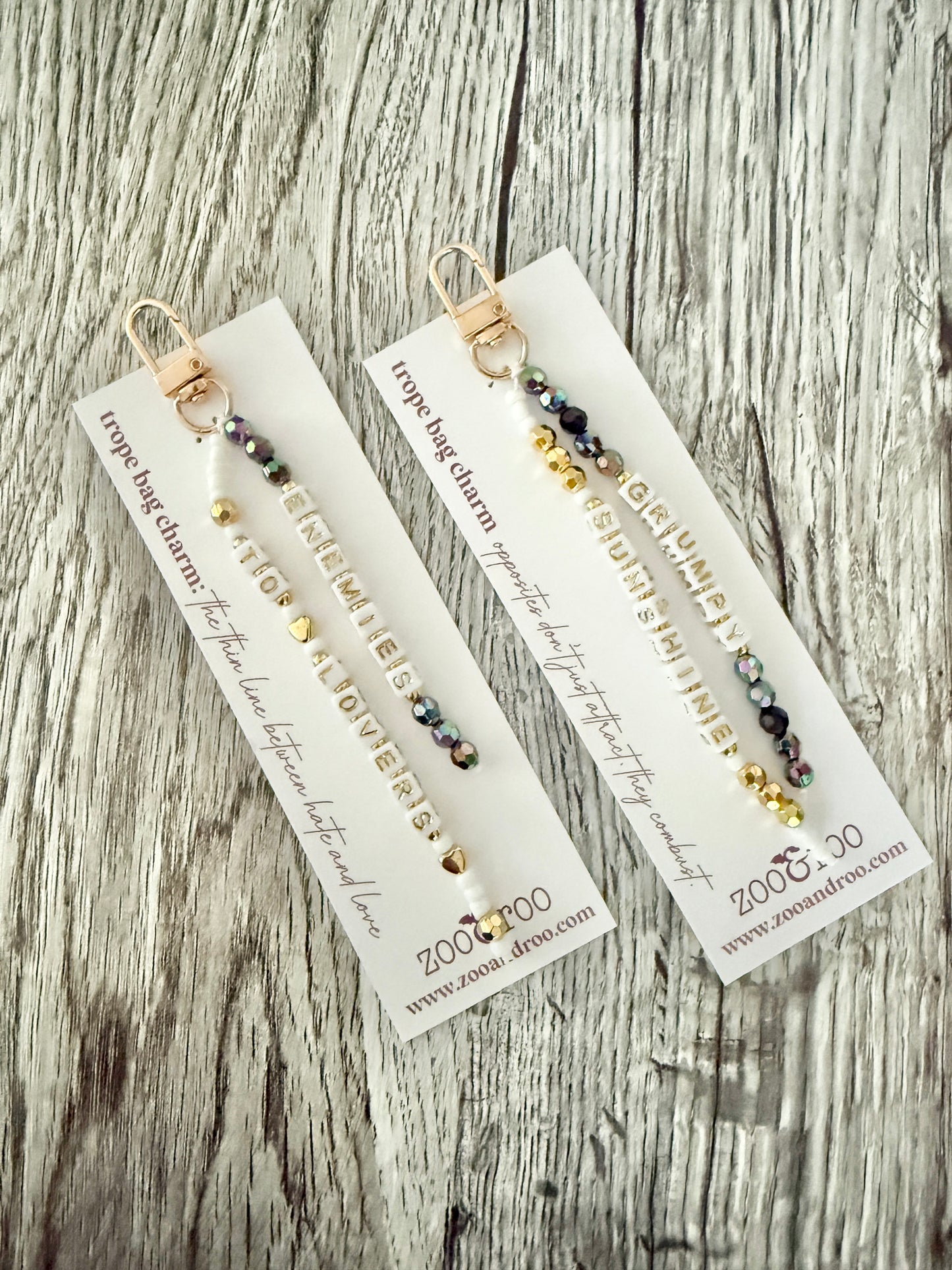 Two gold-toned bag charms with iridescent black, white and gold beads with letter beads that spell 'grumpy/sunshine' and 'enemies to lovers,' displayed on white branded zoo&roo cards over a weathered wood surface.