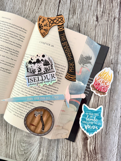 Decorative stickers inspired by Road of Bones by Demi Winters, along with a sword and axe bookmark, on an open book with a wooden background.