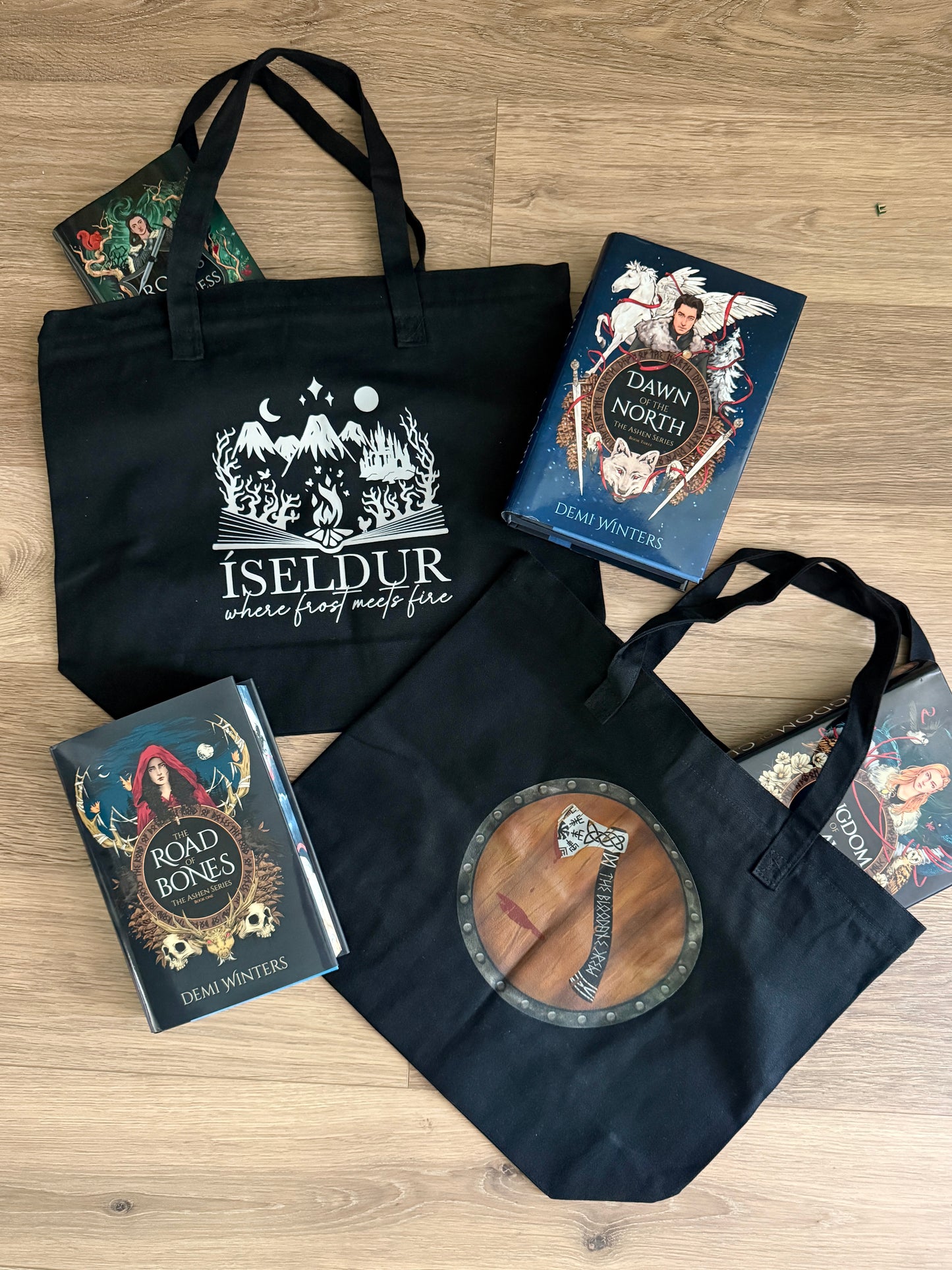 Black tote bags with art inspired by The Road of Bones by Demi Winters, including a bloody axe on a shield and Iseldur spilling from the pages of a book, surrounded by the books from Ashen series.