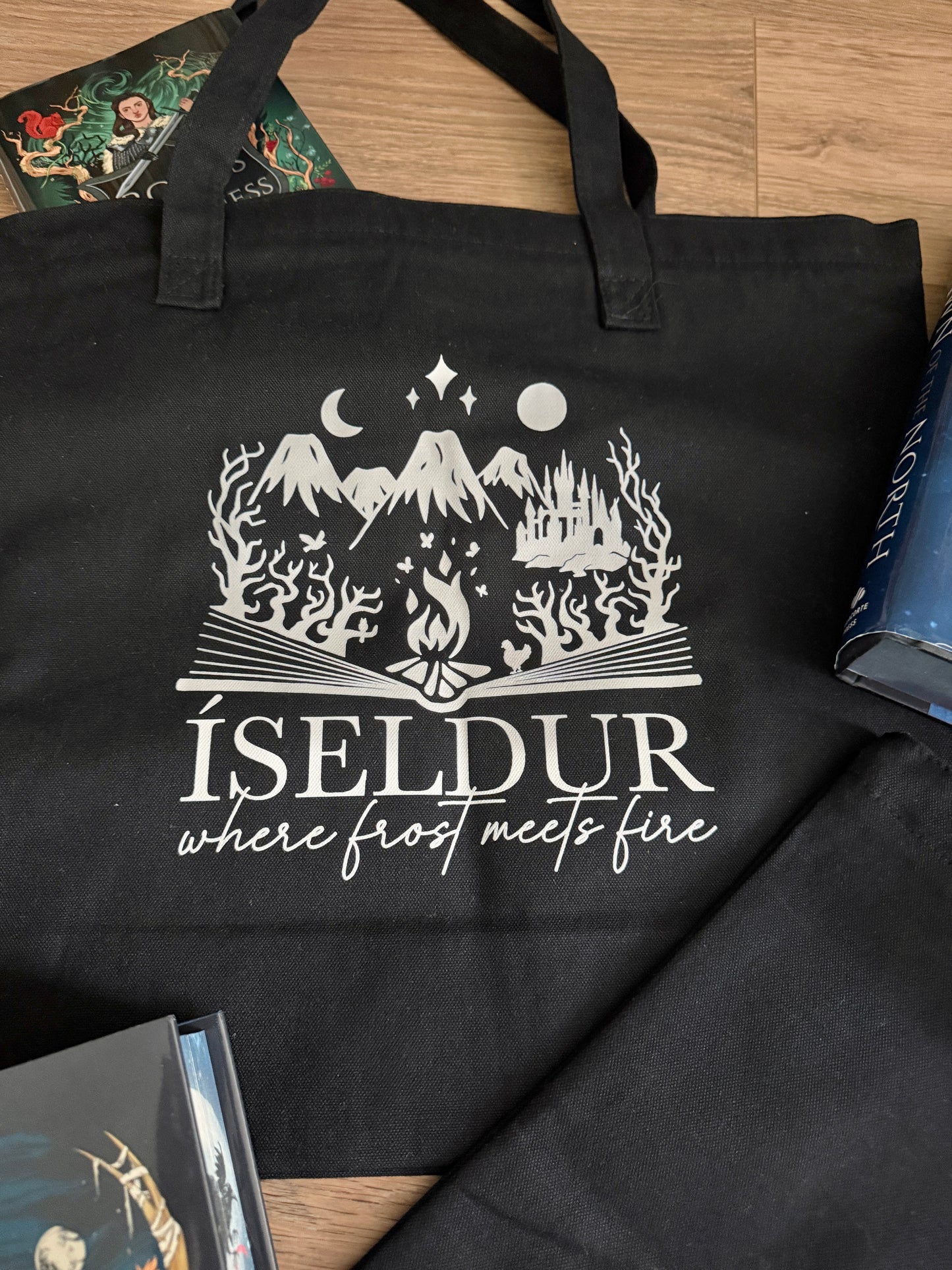 Black tote bag with gray graphic and text 'ÍSELDUR: where frost meets fire' on a wooden surface, inspired by Demi Winters' Ashen series.
