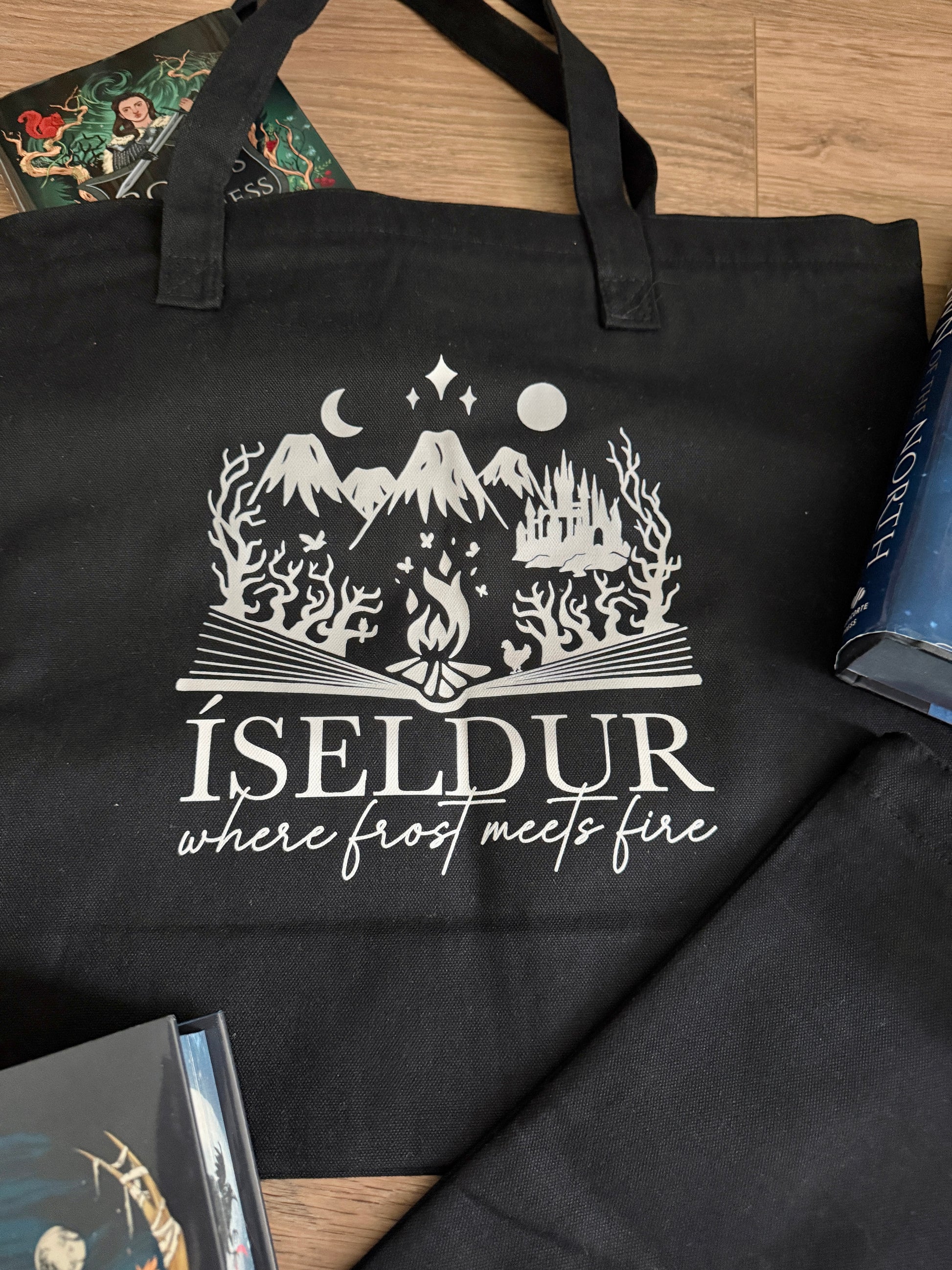Black tote bag with gray graphic and text 'ÍSELDUR: where frost meets fire' on a wooden surface, inspired by Demi Winters' Ashen series.