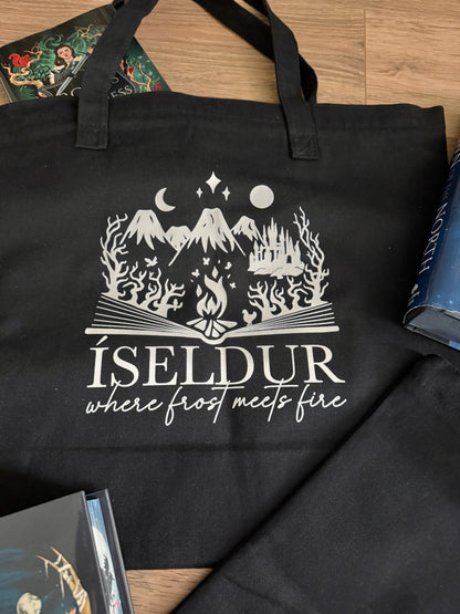 Black tote bag with gray graphic and text 'ÍSELDUR: where frost meets fire' on a wooden surface, inspired by Demi Winters' Ashen series.