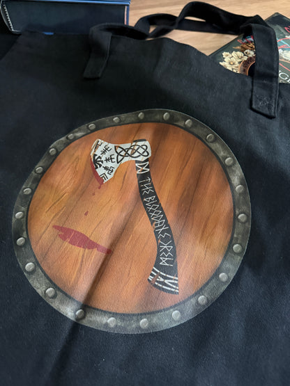 Black tote bag with an axe design on a round wooden shield and The Bloodaxe Crew written in runes, inspired by The Road of Bones by Demi Winters.