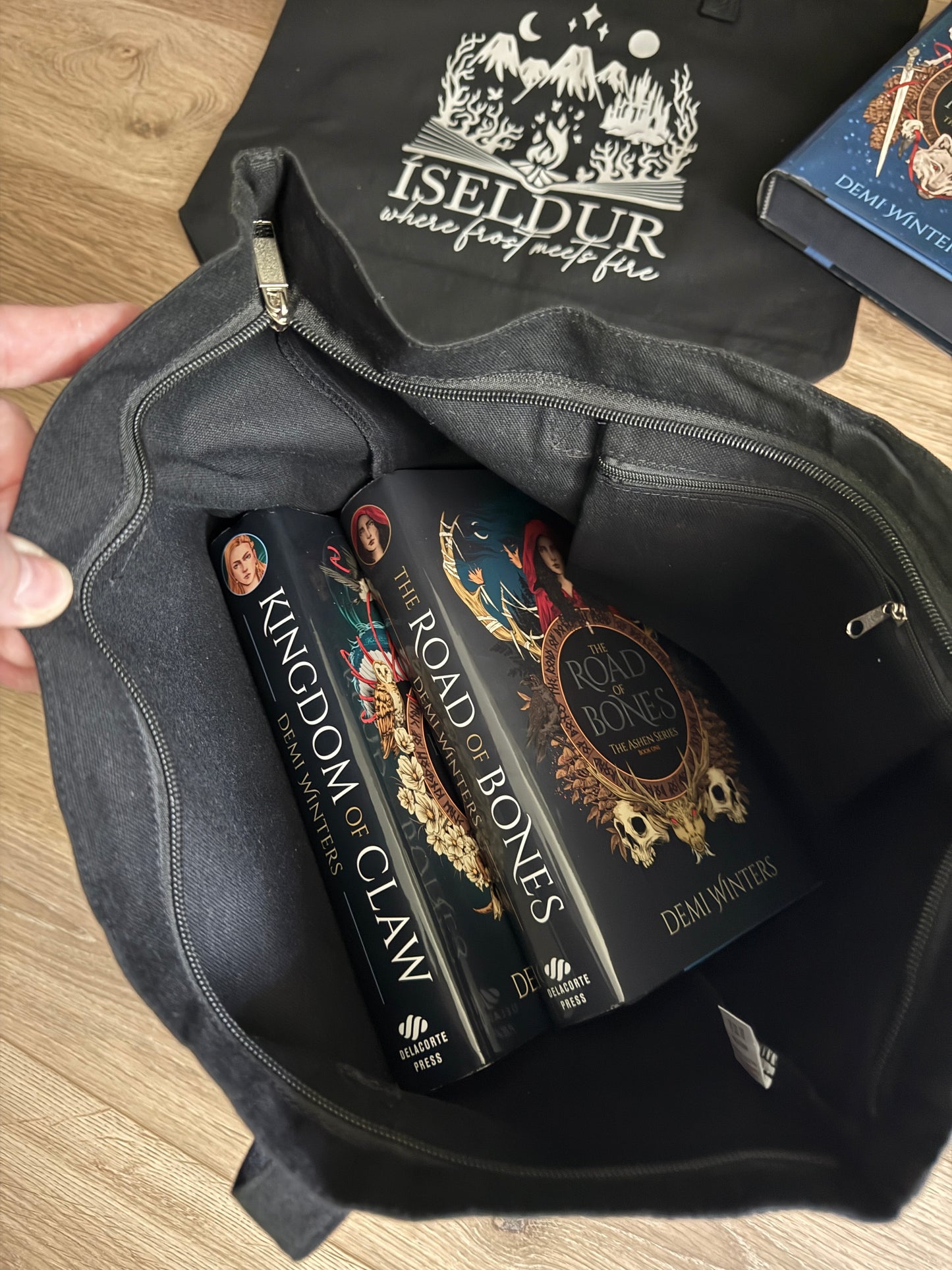 Black tote bag with books inside titled 'Kingdom of Claw' and 'Road of Bones' by Demi Winters, with another black tote bag featuring 'Iseldur: where frost meets fire' branding in the background.