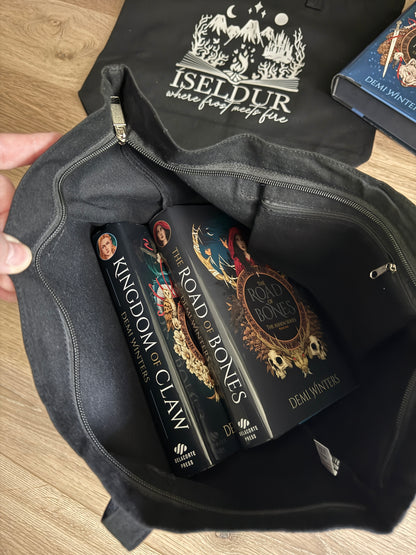 Black tote bag with books inside titled 'Kingdom of Claw' and 'Road of Bones' by Demi Winters, with another black tote bag featuring 'Iseldur: where frost meets fire' branding in the background.