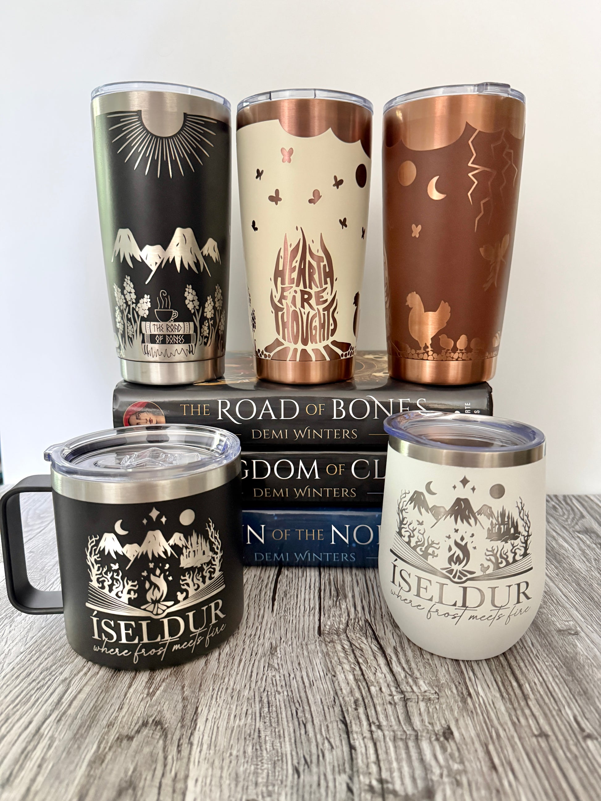 Set of Ashen series tumblers with designs inspired by Demi Winters' Road of Bones on a set of books on a wooden surface