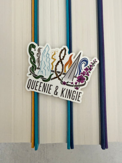 A sticker of a nine pointed crown featuring all the realms of Emarion from the Kindred's Curse Saga books by Penn Cole, with the text 'Queenie & Kingie' laying atop colorful books.
