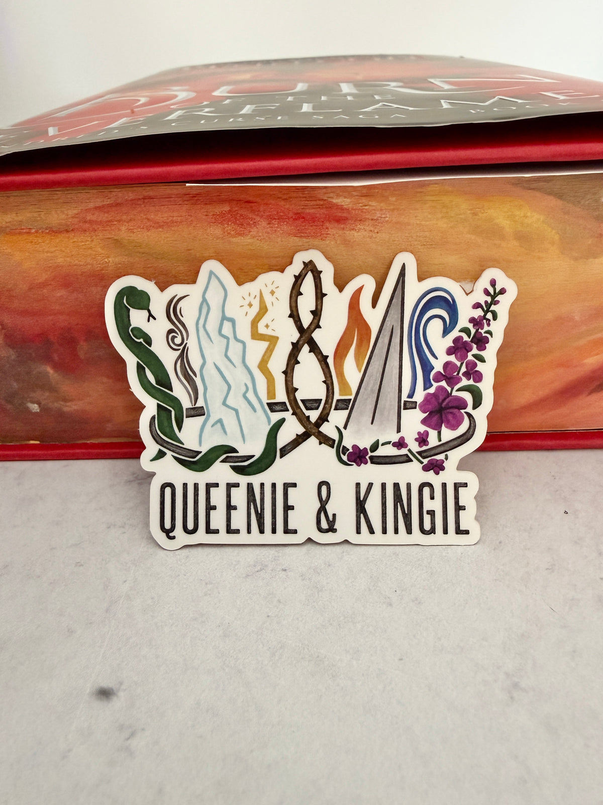 A sticker of a nine pointed crown featuring all the realms of Emarion from the Kindred's Curse Saga books by Penn Cole, with the text 'Queenie & Kingie' leaning against a copy of Burn of the Everflame with edges painted like flames.