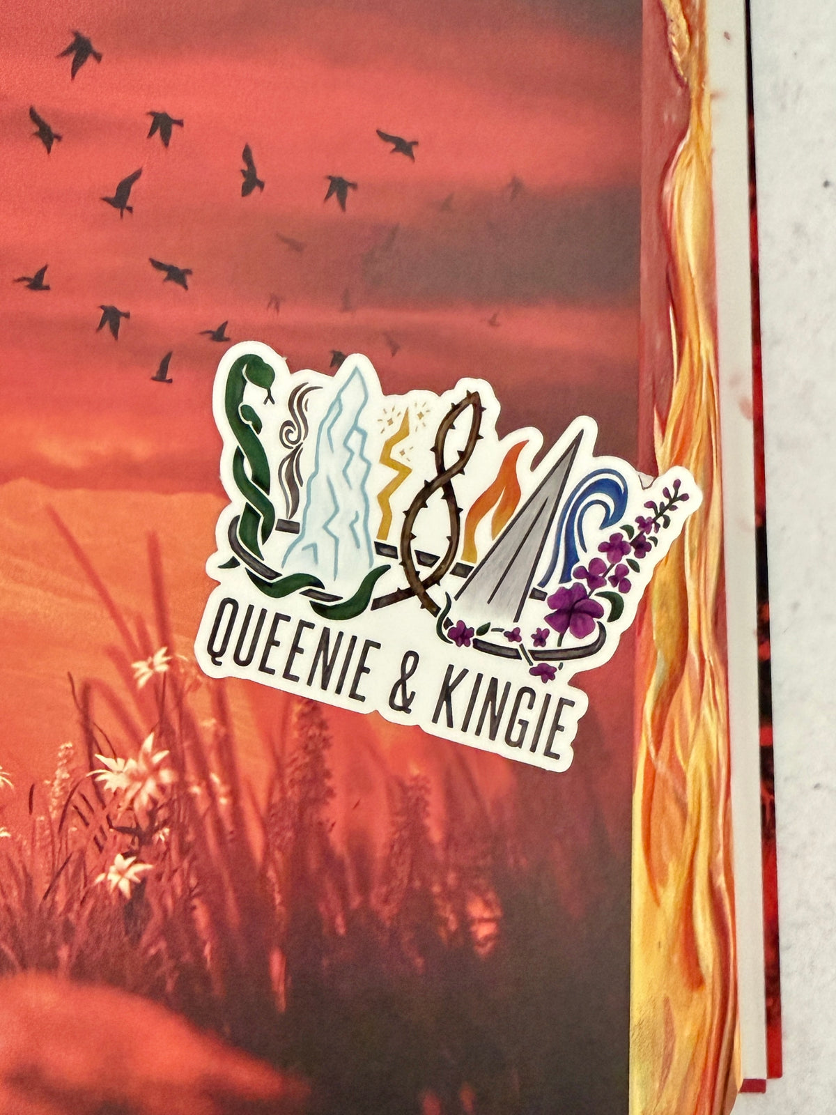 A sticker of a nine pointed crown featuring all the realms of Emarion from the Kindred's Curse Saga books by Penn Cole, with the text 'Queenie & Kingie' laying atop a red hued image of grass and birds.