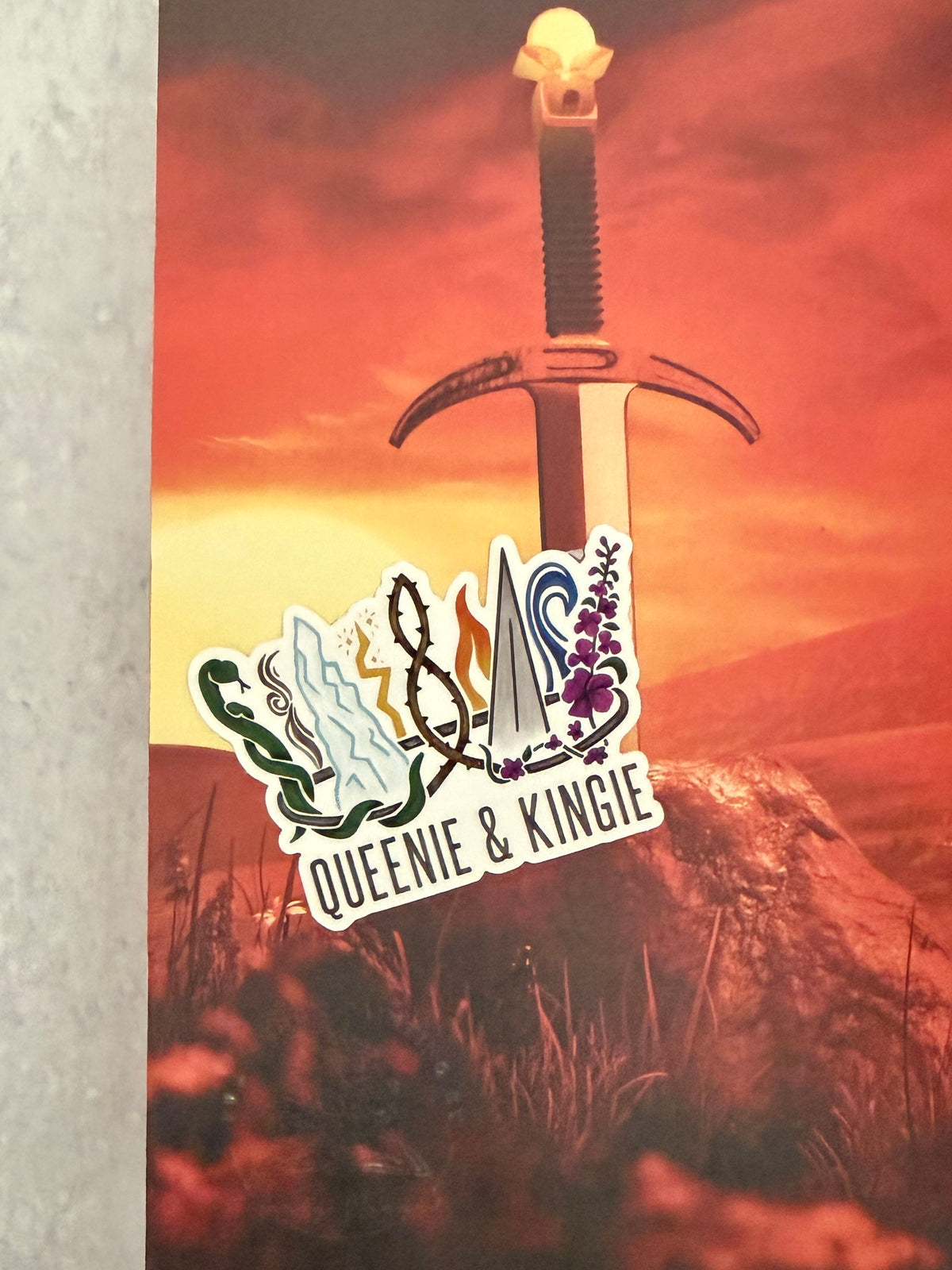 A sticker of a nine pointed crown featuring all the realms of Emarion from the Kindred's Curse Saga books by Penn Cole, with the text 'Queenie & Kingie' laying atop a red hued image of a sword.