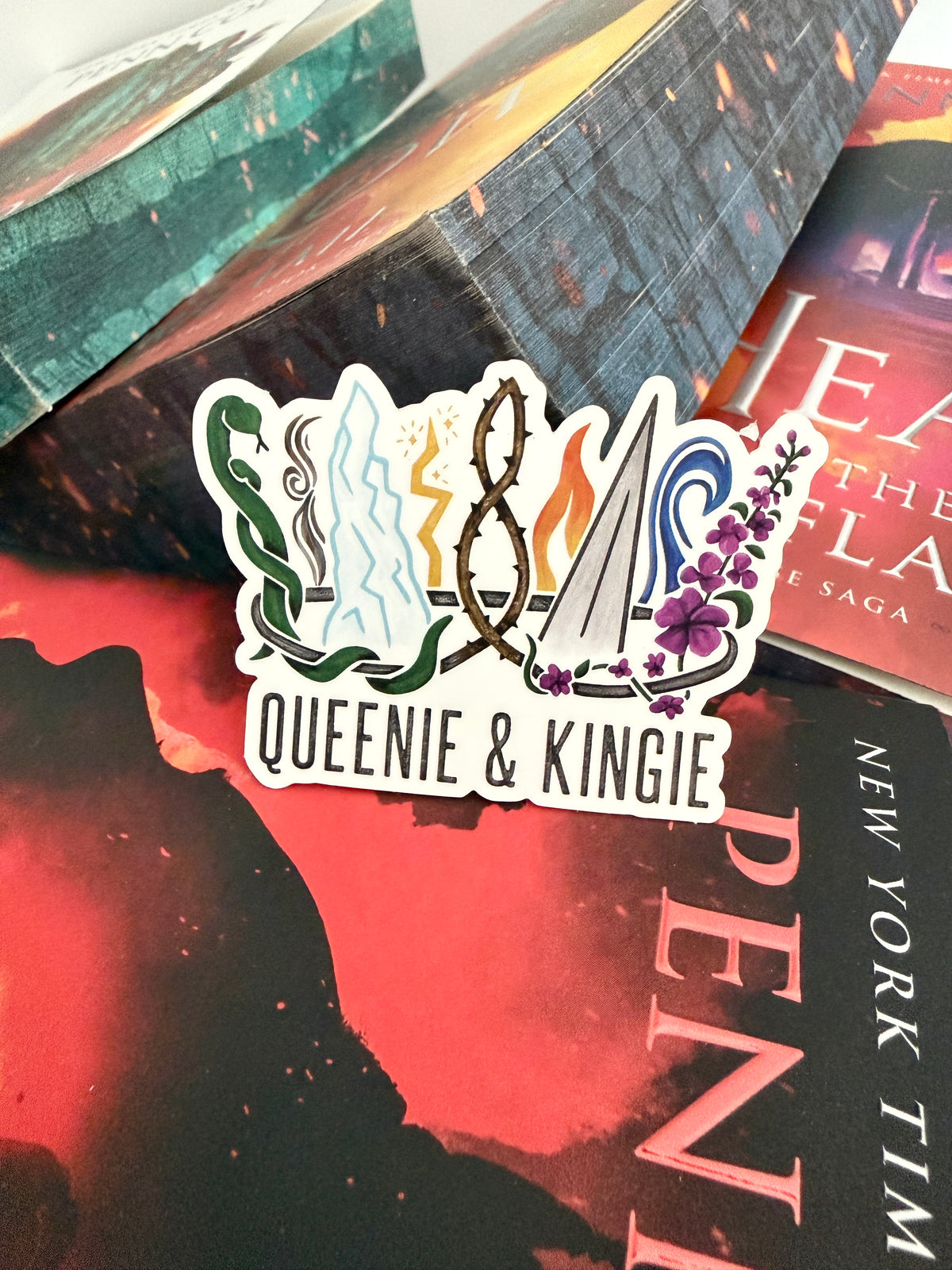 A sticker of a nine pointed crown featuring all the realms of Emarion from the Kindred's Curse Saga books by Penn Cole, with the text 'Queenie & Kingie' laying atop books from the series.