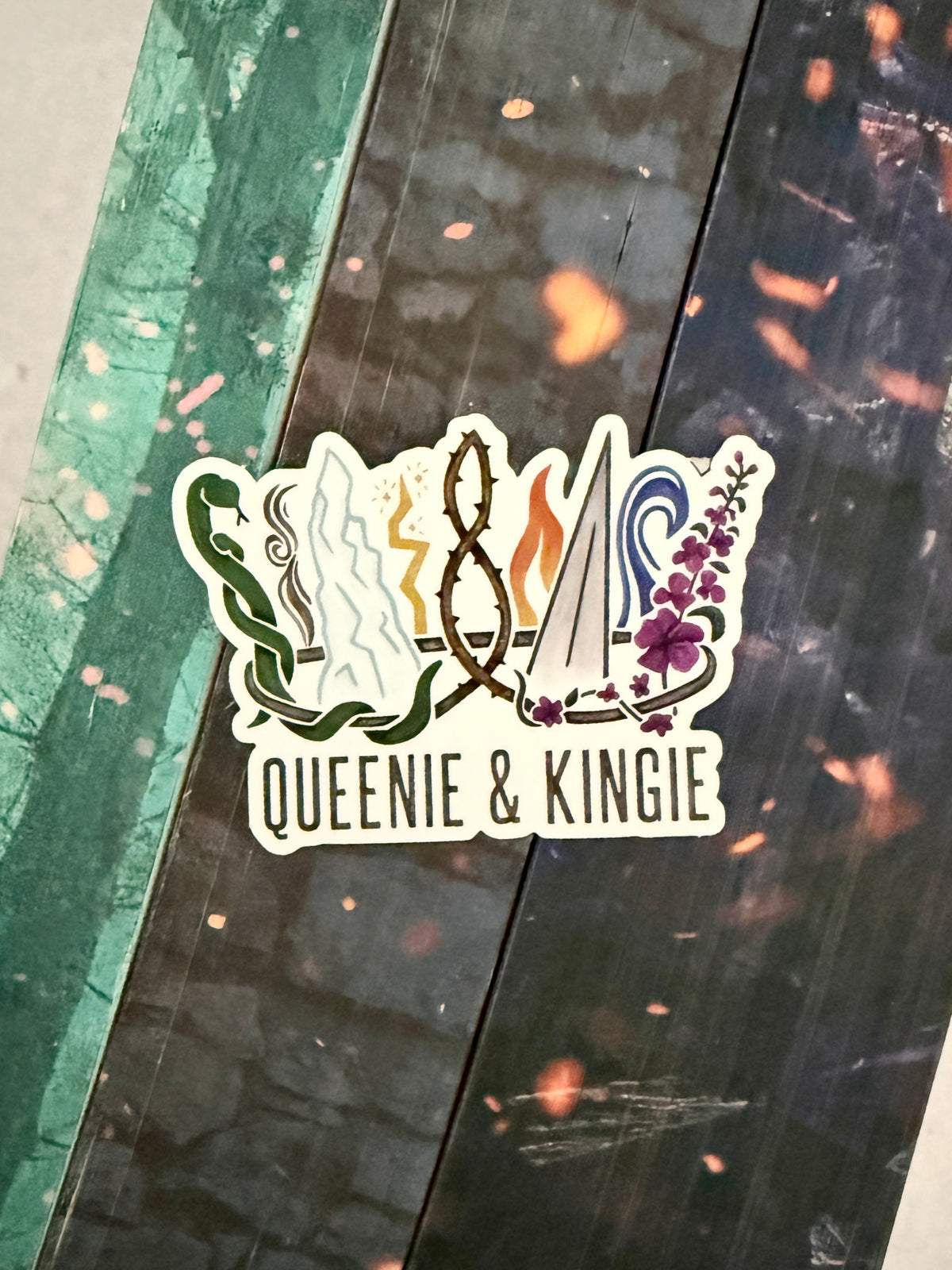 A sticker of a nine pointed crown featuring all the realms of Emarion from the Kindred's Curse Saga books by Penn Cole, with the text 'Queenie & Kingie' laying atop books with edges painted with embers.