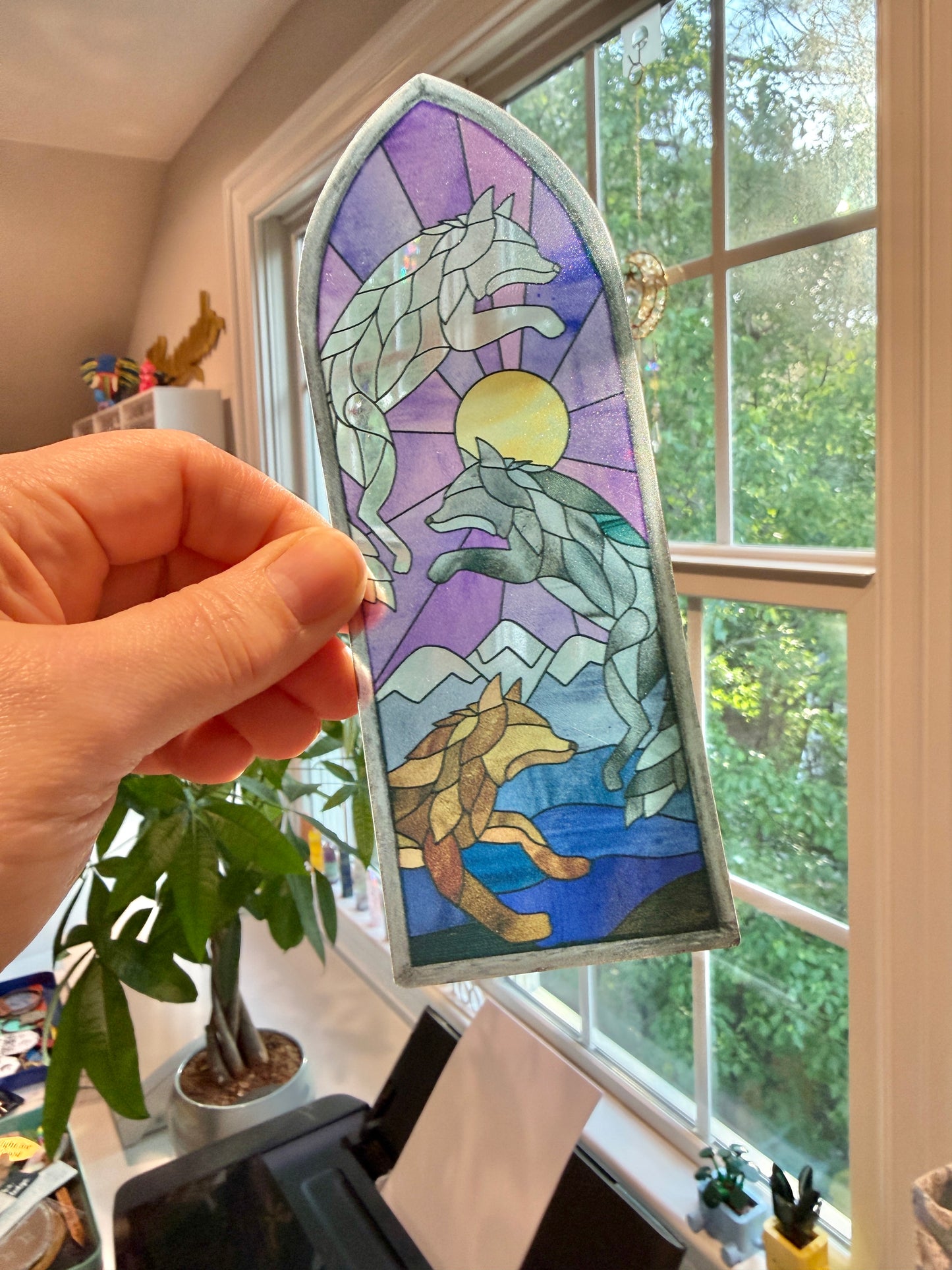 Hand holding a wolf-themed stained glass window bookmark inspired by Dire Bound in front of a window with greenery outside.