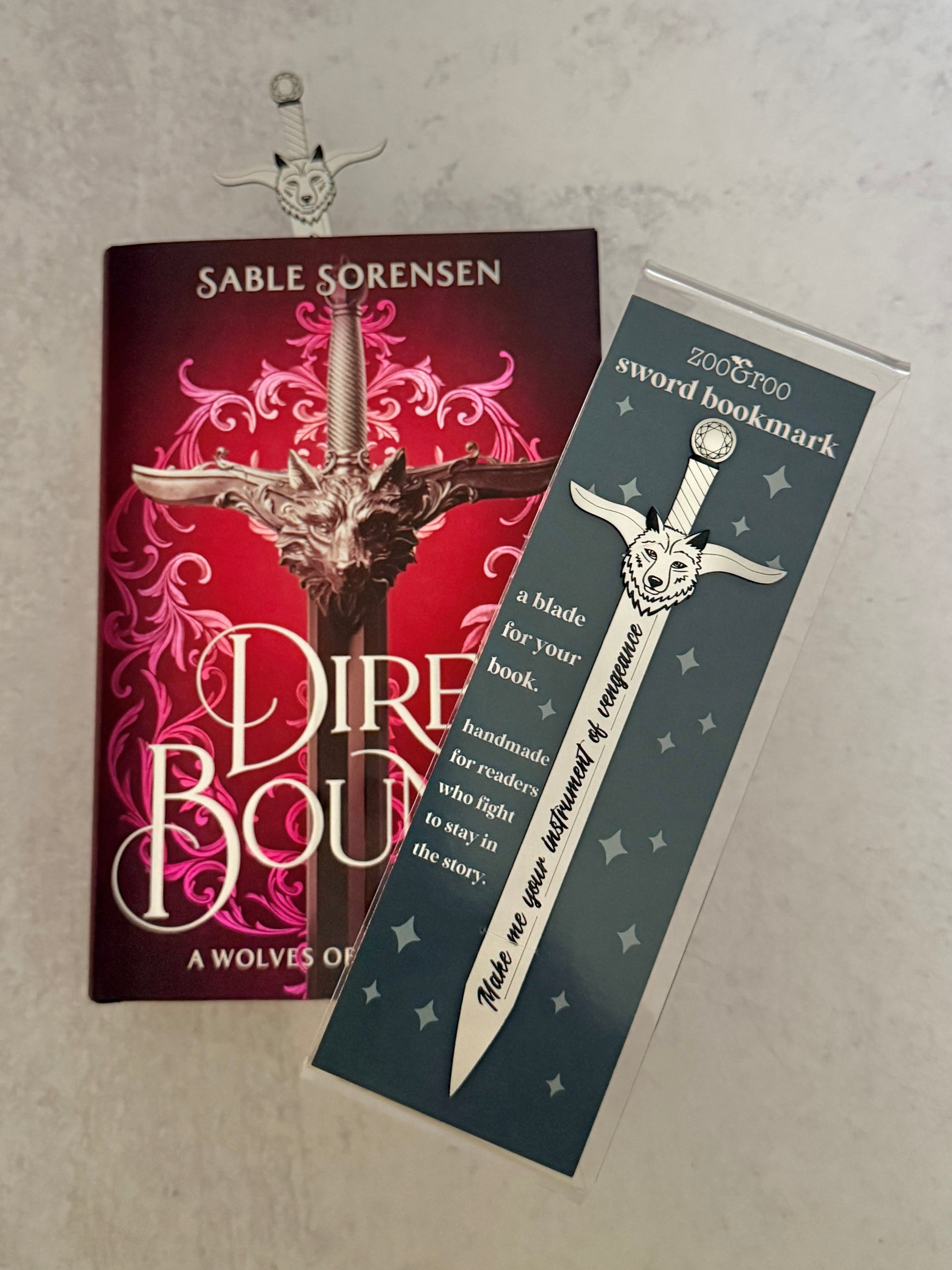 Book 'Dire Bound' by Sable Sorensen with a sword-shaped bookmark engraved with "make me your instrument of vengeance" on a gray background