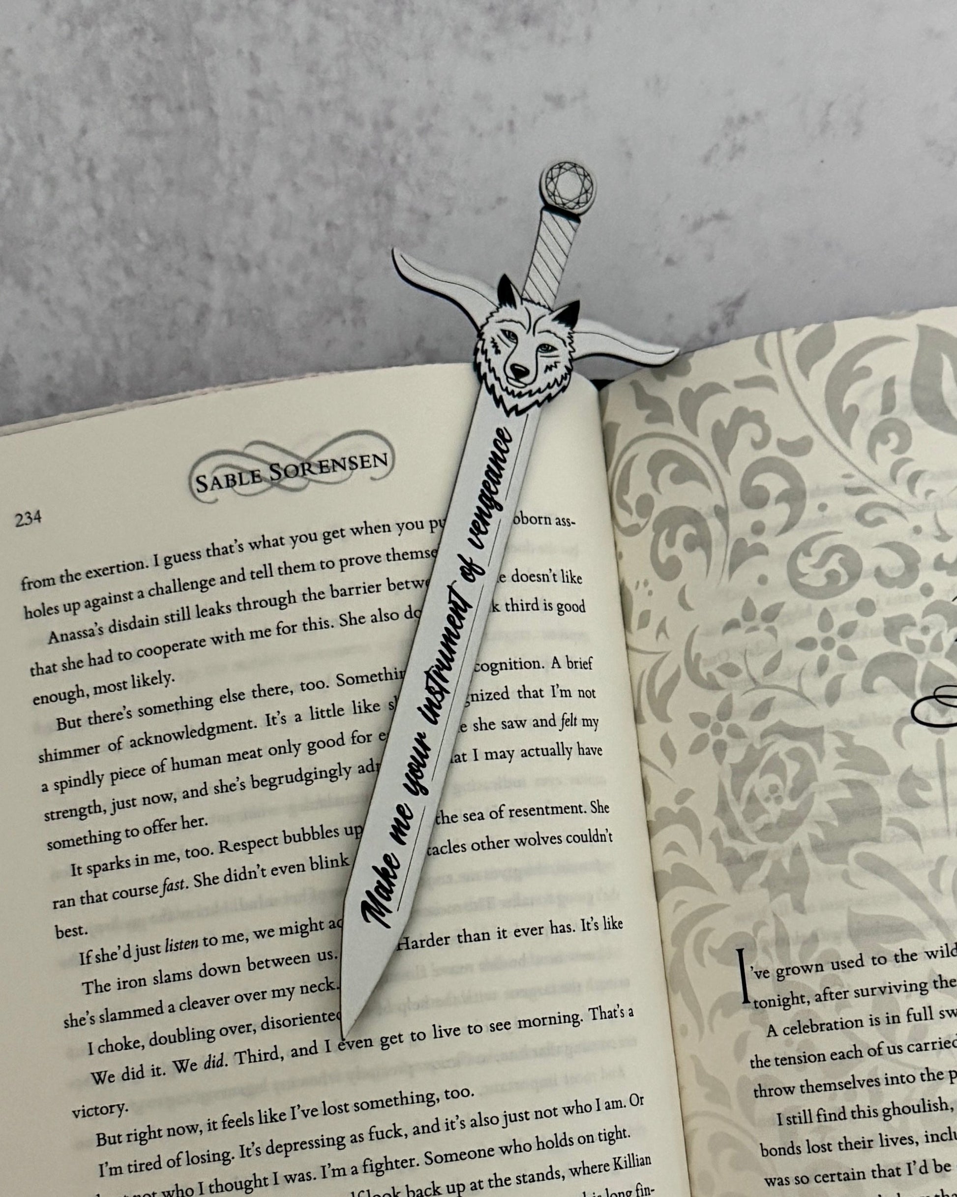 Sword-shaped bookmark with wolf design on an open Dire Bound book by Sable Sorensen engraved with the quote "make me your instrument of vengeance"