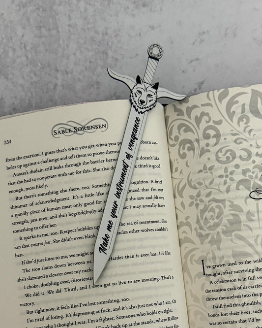Sword-shaped bookmark with wolf design on an open Dire Bound book by Sable Sorensen engraved with the quote "make me your instrument of vengeance"
