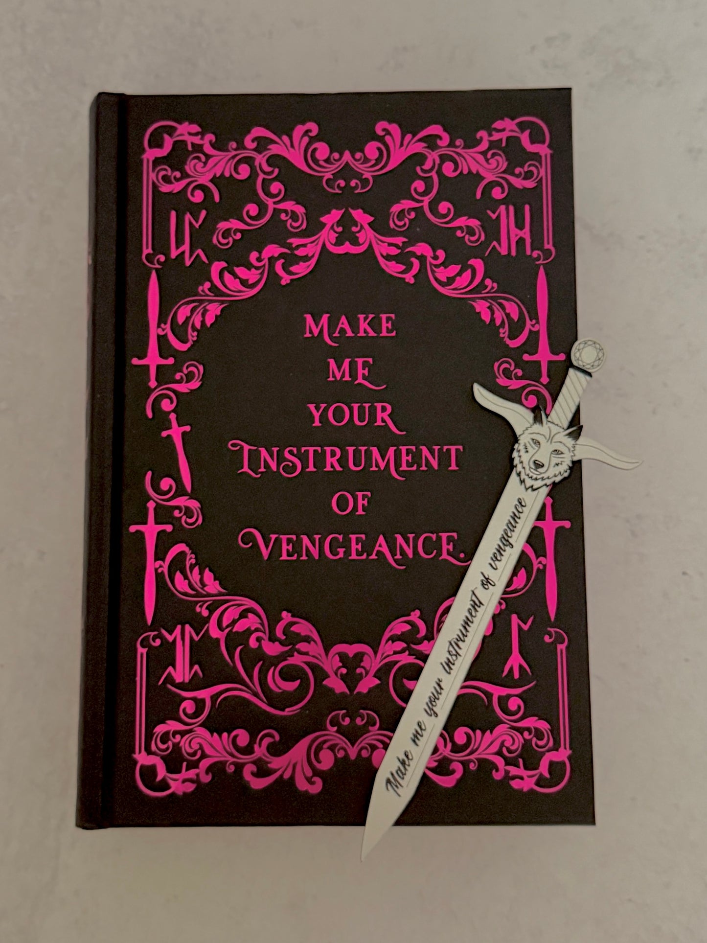 Book with pink decorative text and a white sword bookmark on a gray background - "make me your instrument of vengeance."