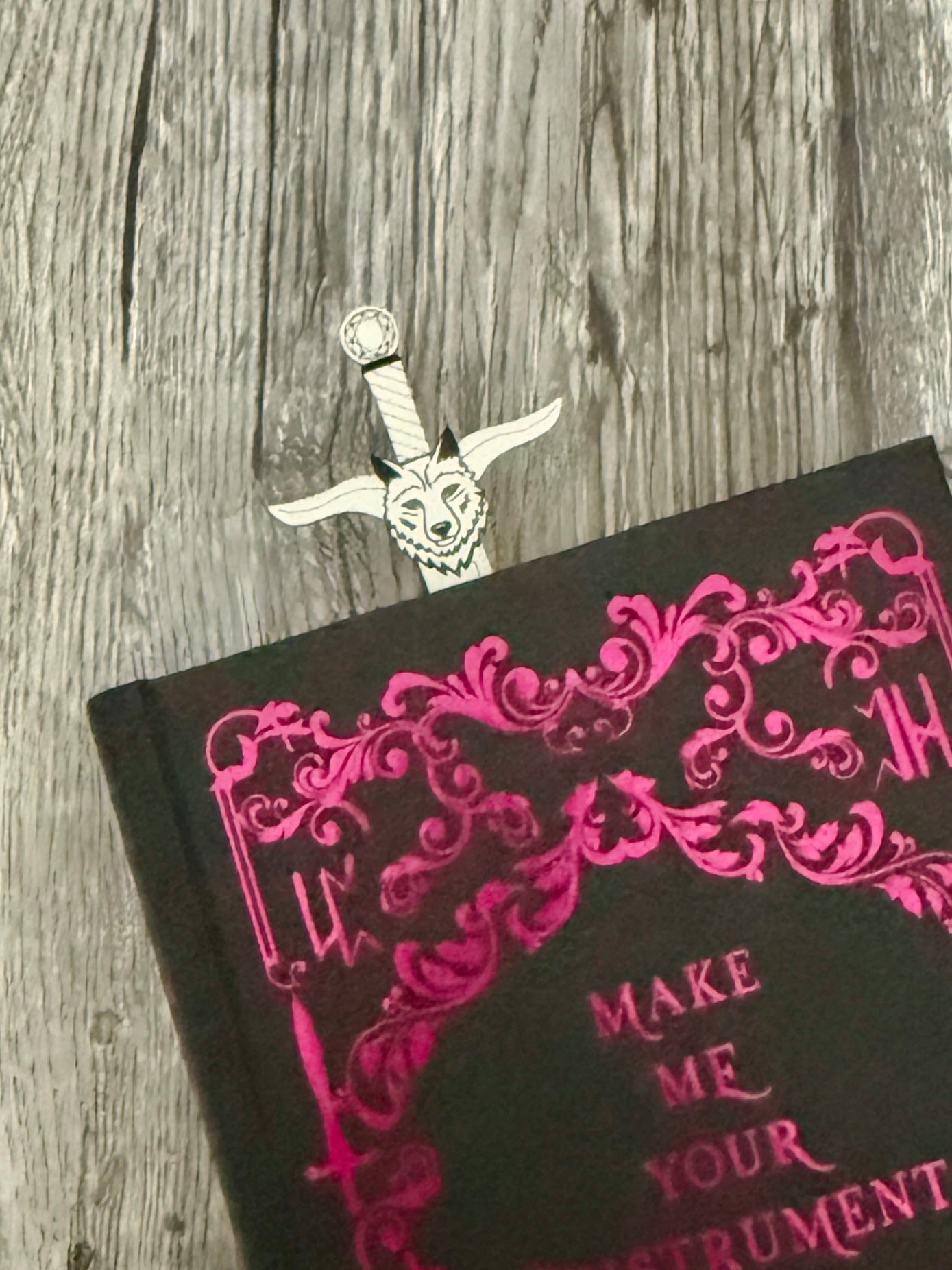 Wolf head pommel of a sword bookmark sticking out of a black book with pink text and decorative design on a wooden surface.