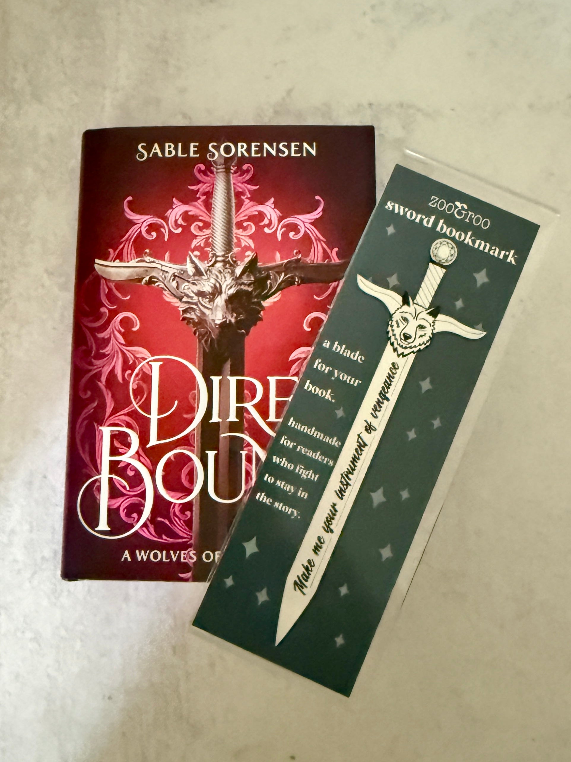 Book titled 'Dire Bound' by Sable Sorensen with a sword bookmark featuring the quote "make me your instrument of vengeance" on a textured surface