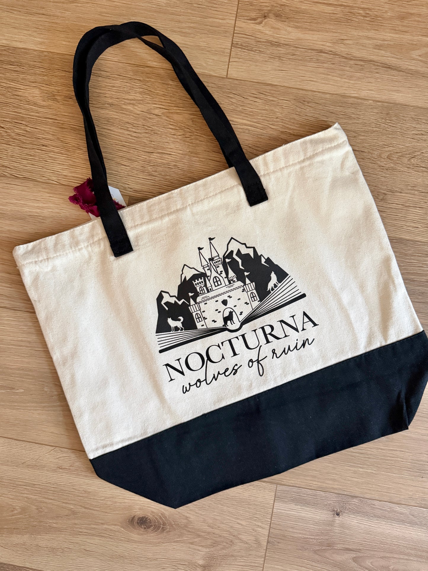Beige tote bag with black handles and Nocturna Wolves of Ruin design inspired by Dire Bound by Sable Sorensen on a wooden surface