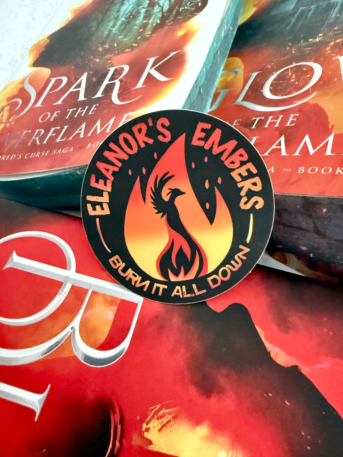 A round black sticker featuring a red and orange phoenix logo with the text 'ELEANOR'S EMBERS' and 'BURN IT ALL DOWN' is placed on top of The Kindred's Curse Saga book covers with matching fiery red and orange artwork.