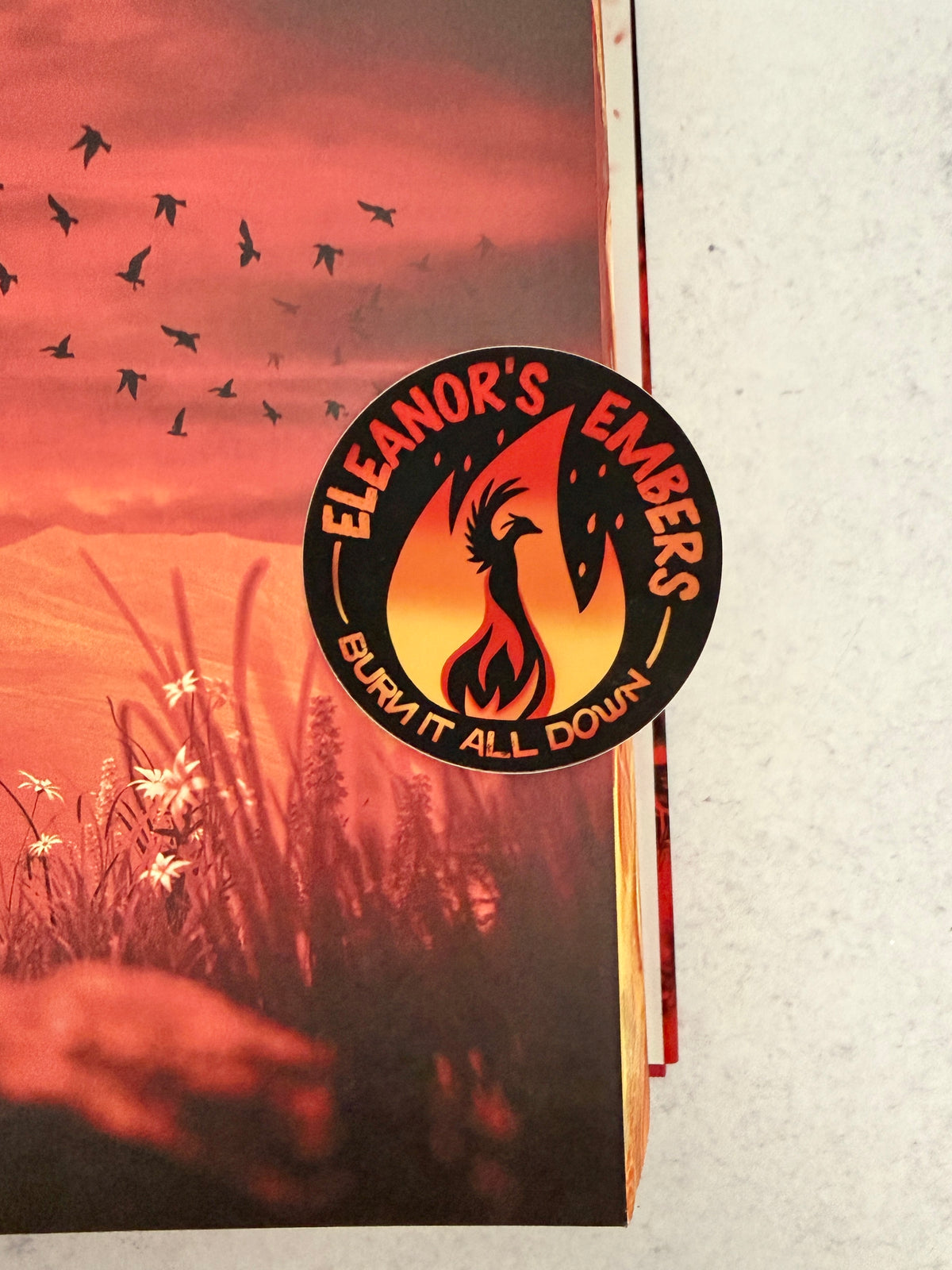 Round black Eleanor's Embers sticker with an orange-to-yellow gradient flame and a black phoenix silhouette, leaning against a fiery image from inside Burn of the Everflame.