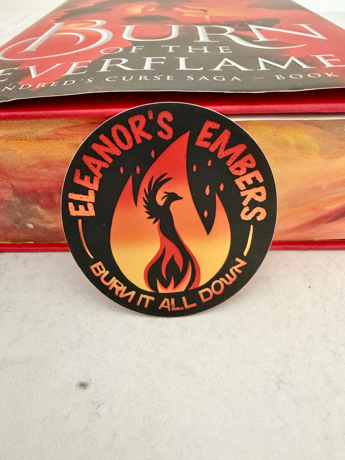 Round black Eleanor's Embers sticker with an orange-to-yellow gradient flame and a black phoenix silhouette, leaning against a copy of Burn of the Everflame.