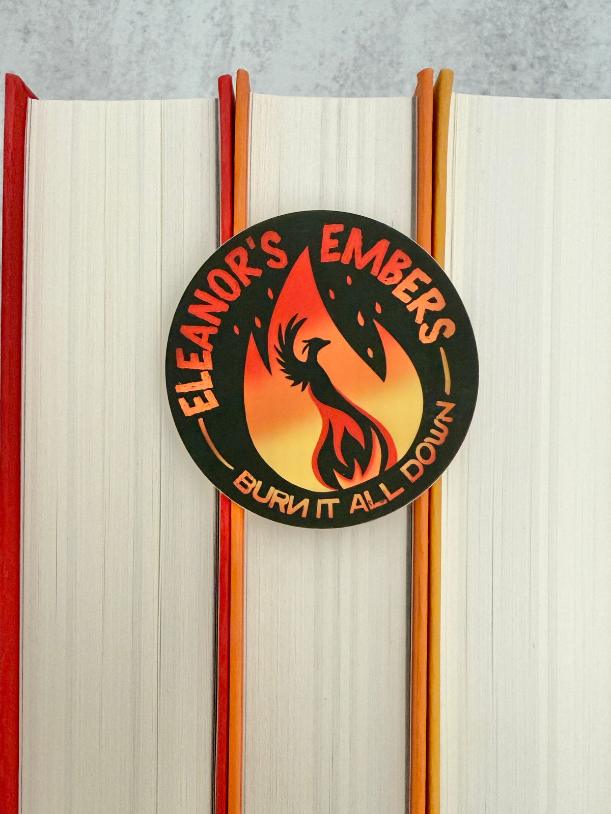 Round black Eleanor's Embers sticker with an orange-to-yellow gradient flame and a black phoenix silhouette, sitting on top of colorful books.