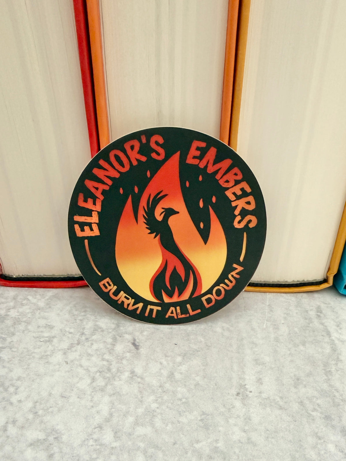 Round black Eleanor's Embers sticker with an orange-to-yellow gradient flame and a black phoenix silhouette, leaning against colorful books.