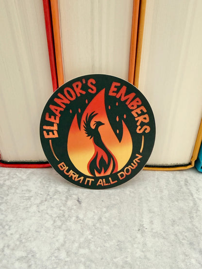 Round black Eleanor's Embers sticker with an orange-to-yellow gradient flame and a black phoenix silhouette, leaning against colorful books.