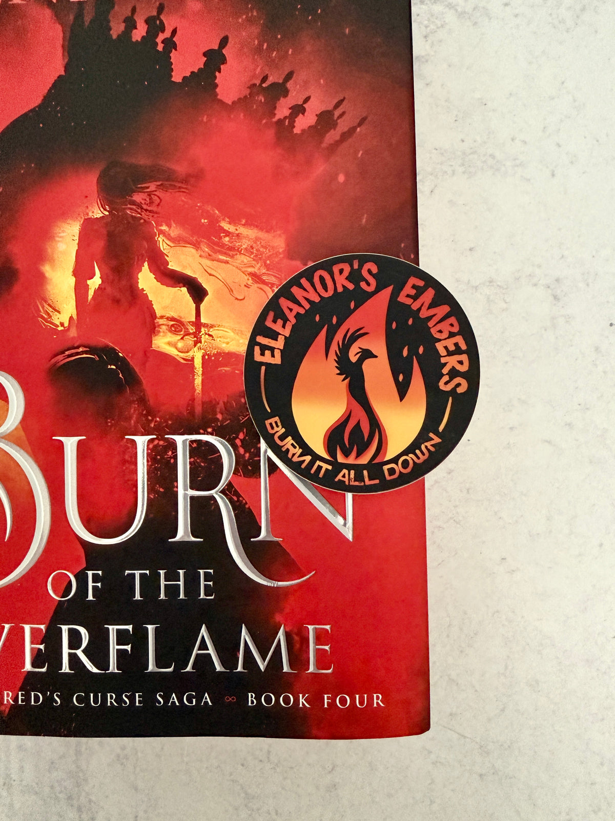 Round black Eleanor's Embers sticker with an orange-to-yellow gradient flame and a black phoenix silhouette, sitting on top of a copy of Burn of the Everflame by Penn Cole.