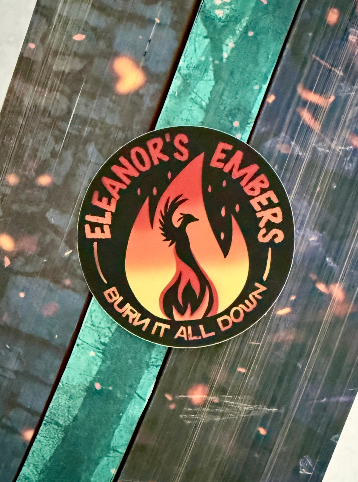 Round black Eleanor's Embers sticker with an orange-to-yellow gradient flame and a black phoenix silhouette, sitting on top of books from the Kindred's Curse Saga by Penn Cole.