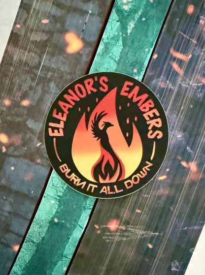 Round black Eleanor's Embers sticker with an orange-to-yellow gradient flame and a black phoenix silhouette, sitting on top of books from the Kindred's Curse Saga by Penn Cole.