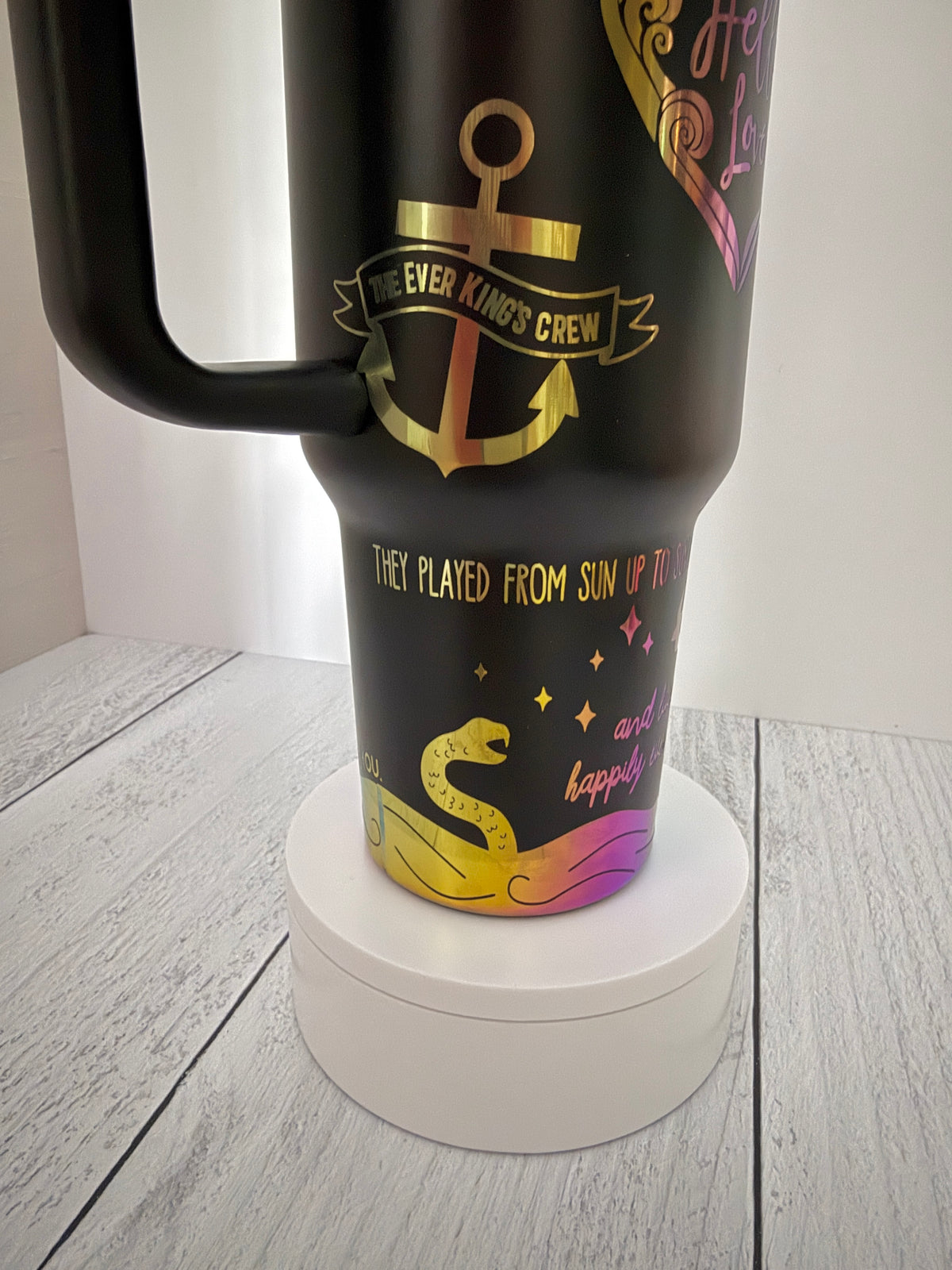 A black insulated Ever King themed tumbler with a handle, featuring rainbow engraved graphics including an anchor, a serpent, and quotes from the book, displayed on a white circular pedestal against a light gray wood floor and white wall.