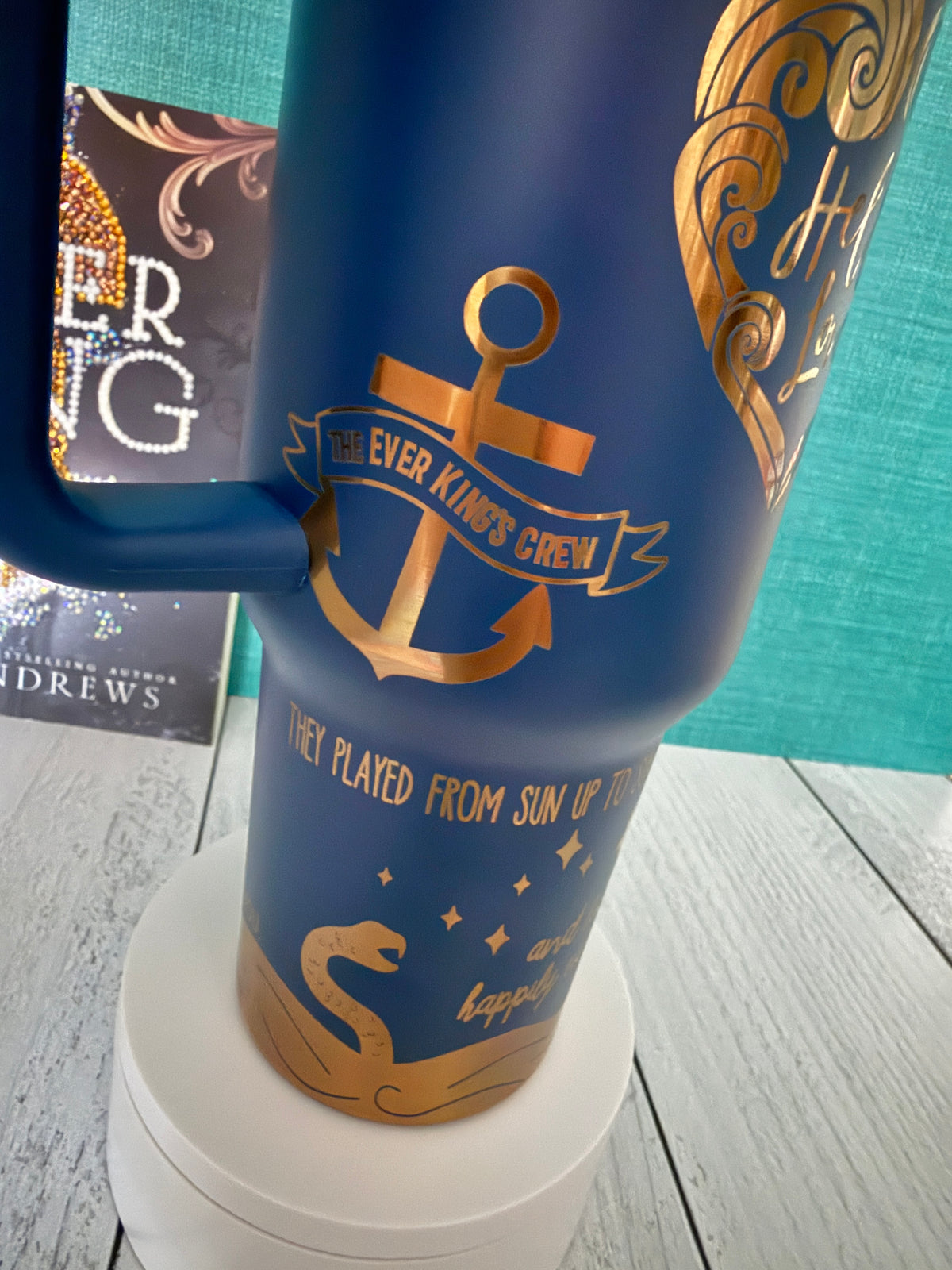 An Ever King themed navy blue insulated tumbler with gold engraved graphics, including an anchor, the text 'The Ever Kings Crew,' and quotes from the book by LJ Andrews, shown on a white pedestal with a teal wall and gray wood floor in the background.