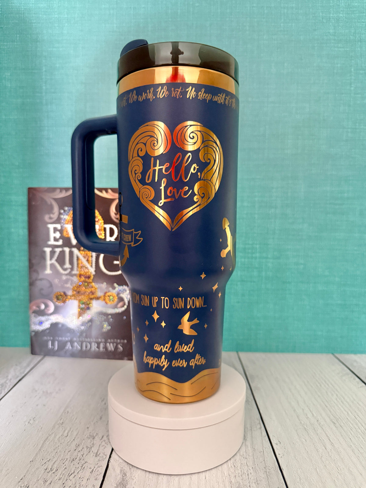 A navy blue insulated travel tumbler with a gold heart design that says 'hello love' and gold text, displayed on a white pedestal. The Ever King book by LJ Andrews is visible in the background against a teal wall.