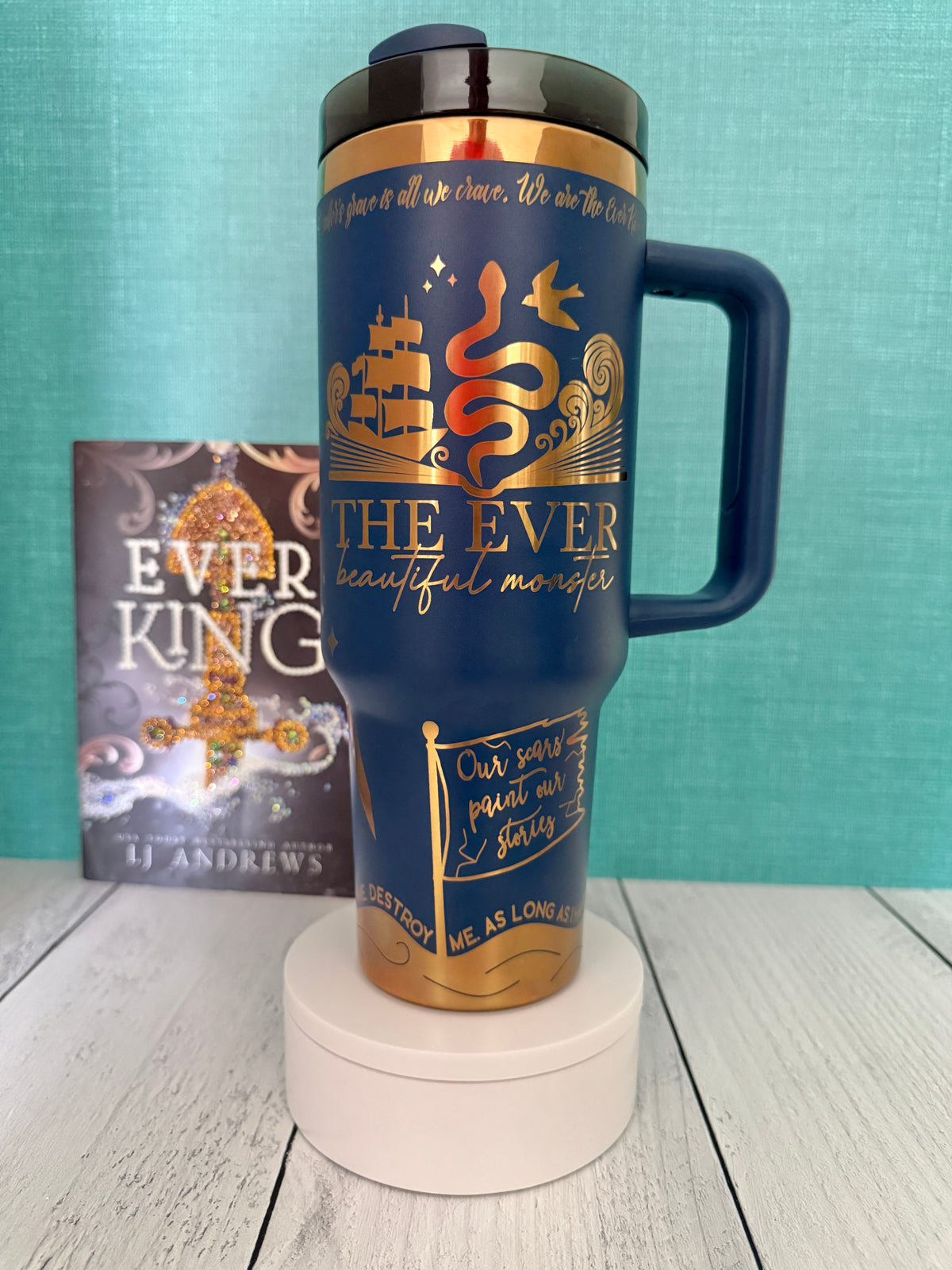 An Ever King themed navy handled travel tumbler sits on a white pedestal in front of a teal wall and light wood floor. The tumbler is printed with the title 'The Ever: Beautiful Monster' in script and serif fonts, decorated with a serpent, ship, songbird and copper engraved details.