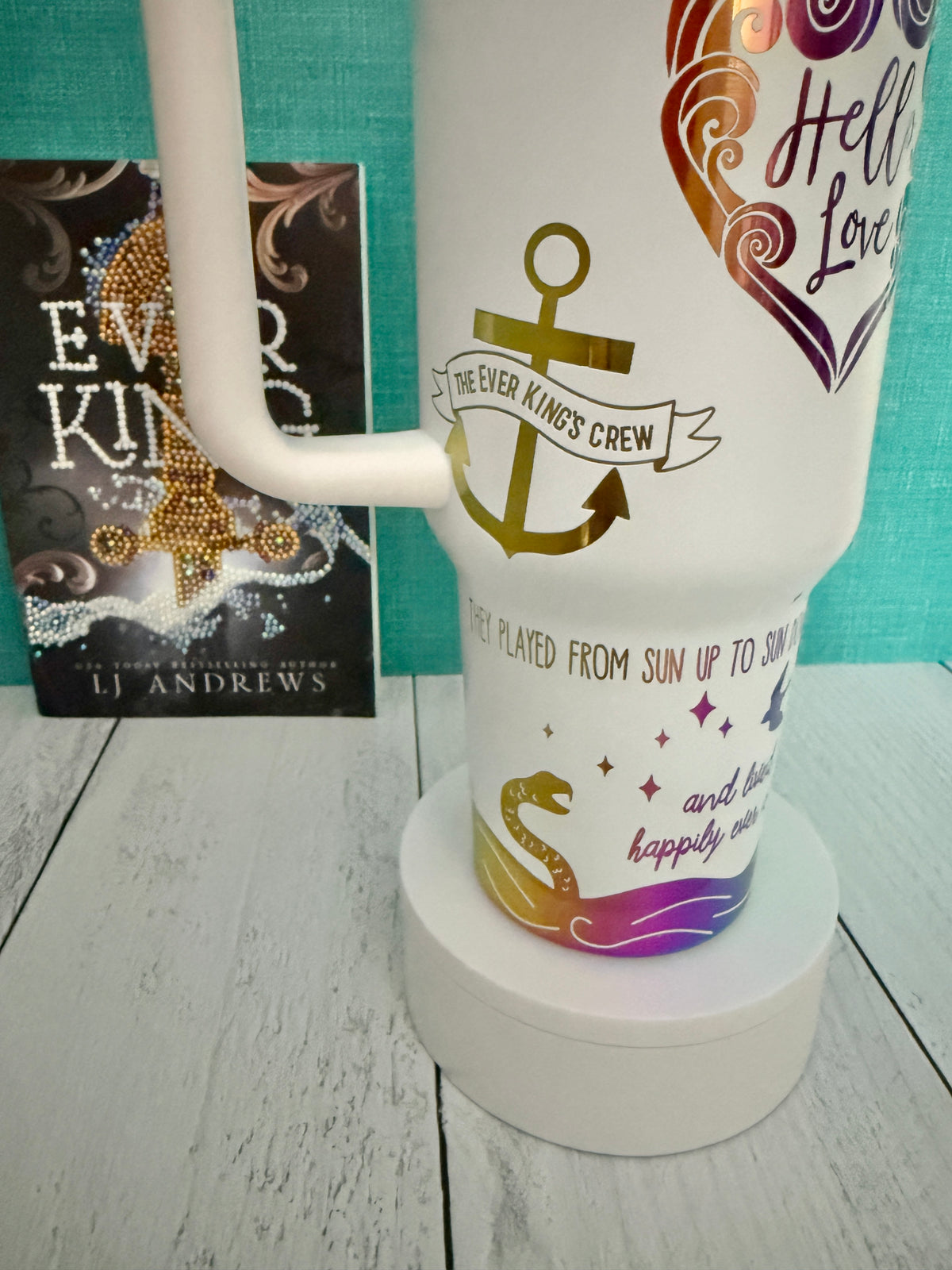 An Ever King themed white handled travel tumbler on a white pedestal in front of a teal wall and light wood floor. The tumbler is engraved with an anchor, waves, serpent and quotes from the book by LJ Andrews.