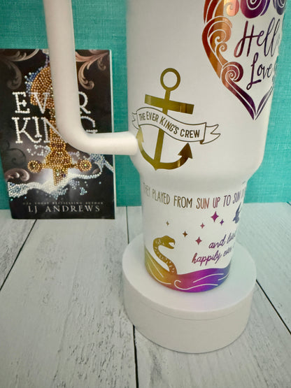 An Ever King themed white handled travel tumbler on a white pedestal in front of a teal wall and light wood floor. The tumbler is engraved with an anchor, waves, serpent and quotes from the book by LJ Andrews.