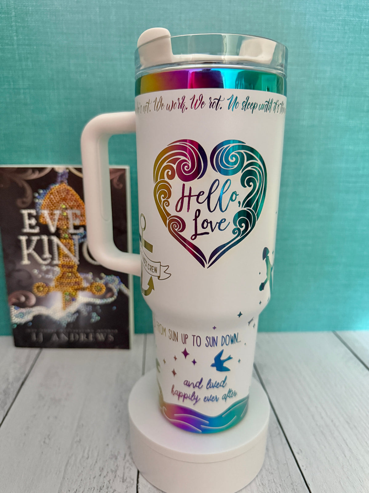 A white insulated travel tumbler with a handle and clear lid, featuring a large iridescent rainbow heart design with the words 'Hello, Love' on the front. The tumbler is displayed on a white pedestal with a teal background and a book titled 'Ever King' by LJ Andrews.
