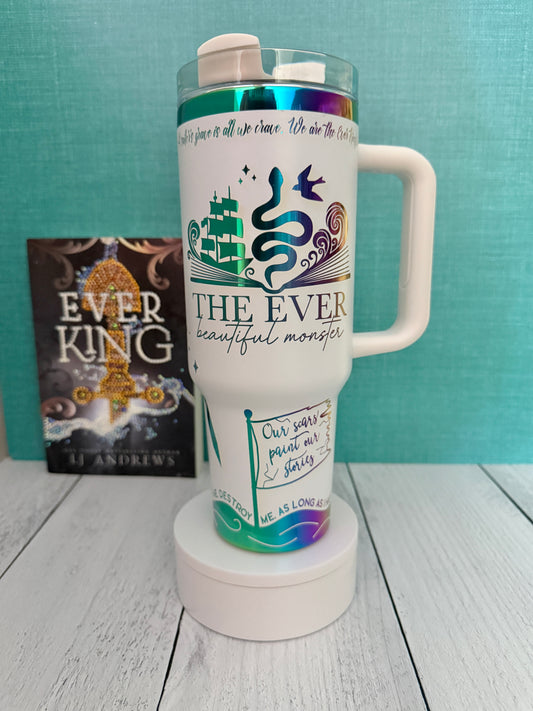 An Ever King themed white handled travel tumbler with a clear lid sits on a white pedestal in front of a teal wall and light wood floor. The tumbler is printed with the title 'The Ever: Beautiful Monster' in script and serif fonts, decorated with a serpent, ship, songbird and rainbow engraved details.