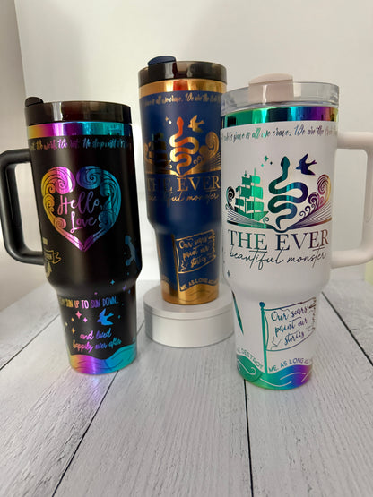Three Ever King themed insulated travel tumblers with handles and lids are displayed on a white wooden surface against a white wall. The left tumbler is black with a holographic rainbow heart design and 'Hello, Love!' text. The right is white with a rainbow engraved 'The Ever: beautiful monster' book design.