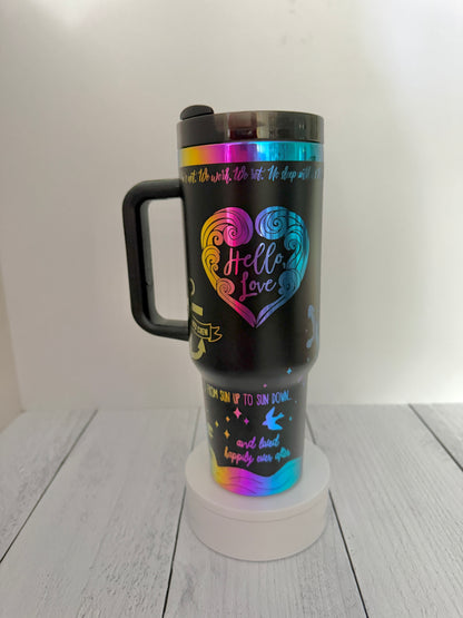 A black handled Ever King themed travel tumbler with a lid, featuring a colorful holographic rainbow heart design and inspirational text from the book, displayed on a white circular stand against a light gray background.
